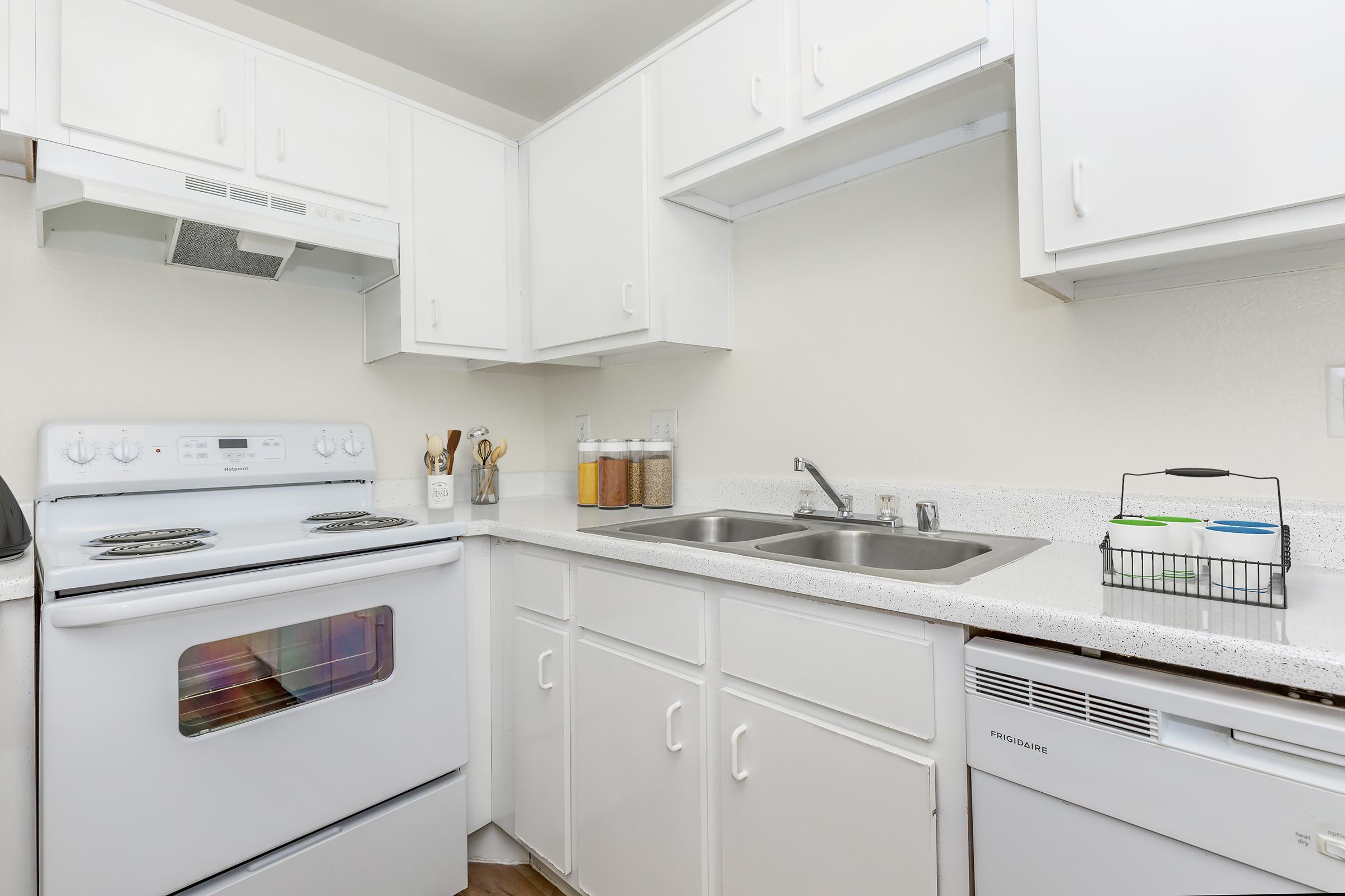 A clean, modern kitchen featuring white cabinetry, a stove with an oven, a double sink, and a dishwasher. Countertops display jars of dry goods, and kitchen utensils are arranged neatly. The overall aesthetic is bright and organized, emphasizing functionality and simplicity.