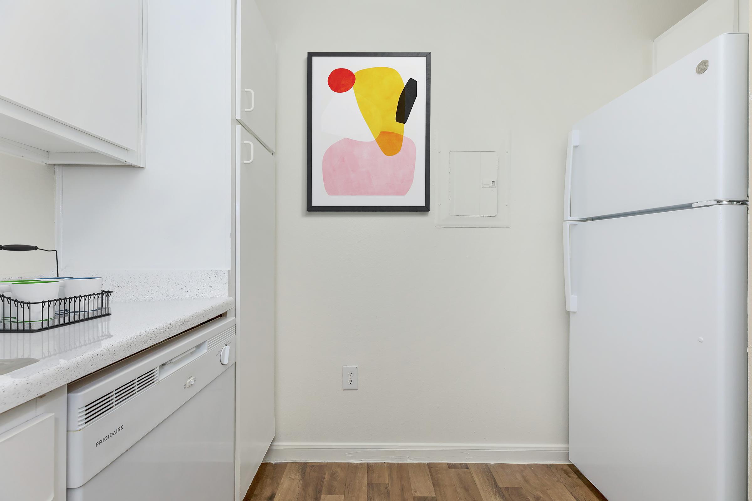 A modern kitchen featuring white cabinets and appliances, including a refrigerator. The wall displays a colorful abstract painting with shapes in red, yellow, and pink. The flooring is a warm wood tone, and there is a small basket on the countertop.