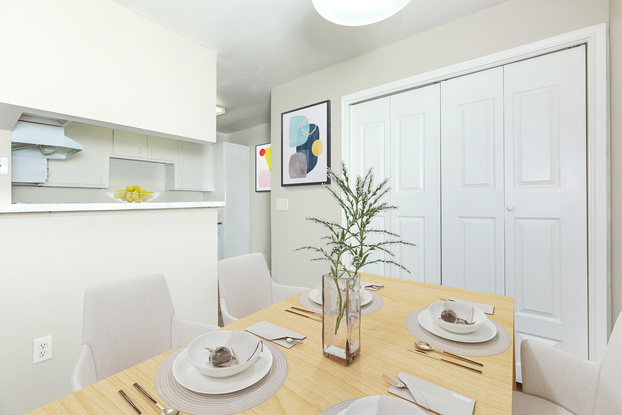 A modern dining area featuring a wooden table set for four with stylish plates and cutlery. A minimalist vase with greenery is the centerpiece. Behind, a well-lit kitchen area is visible, along with a framed abstract art piece on the wall and white double closets. The decor emphasizes simplicity and elegance.