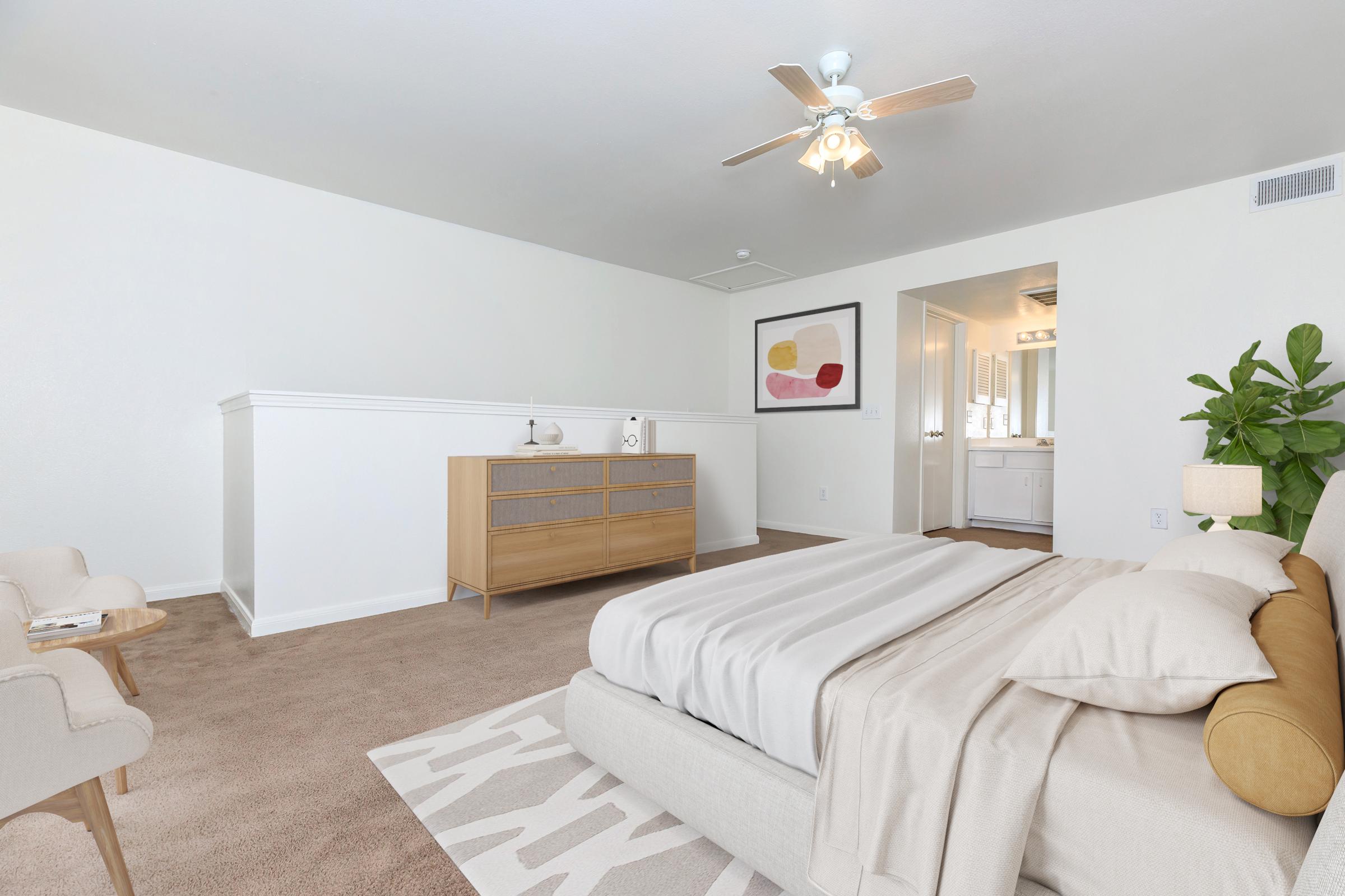 A spacious, well-lit bedroom featuring a neutral color palette. The room includes a beige bed with cozy linens, a wooden dresser, a comfortable armchair, and a ceiling fan. A plant adds a touch of greenery, and a doorway leads to an adjoining bathroom area. Decor includes a framed abstract artwork on the wall.