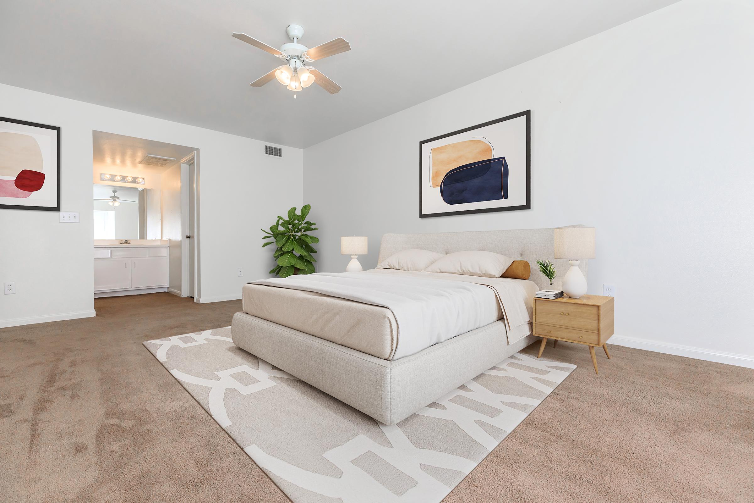 A cozy bedroom featuring a light-colored bed with beige bedding, two bedside lamps, and a wooden nightstand. A large area rug with a geometric pattern lies on the floor. In the background, there's a doorway leading to a bathroom, and a potted plant adds a touch of greenery to the space.
