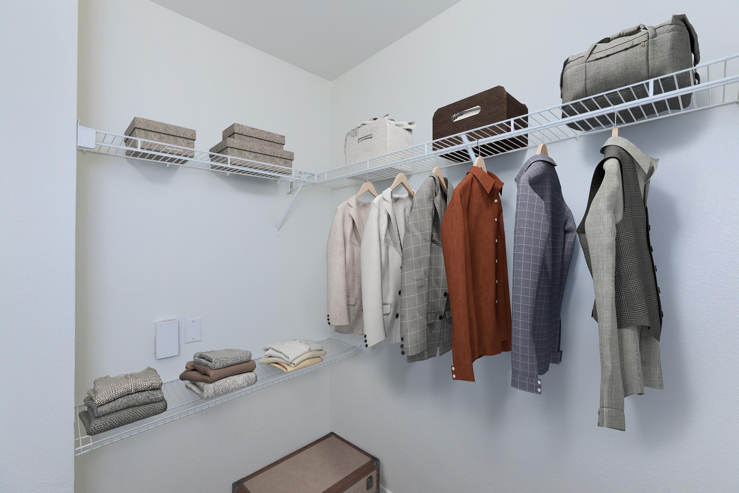 A neatly organized walk-in closet featuring shelves with various storage boxes, folded sweaters, and hanging clothing items in neutral tones, including both patterned and solid shirts. A suitcase is positioned at the bottom, contributing to a tidy and inviting space.