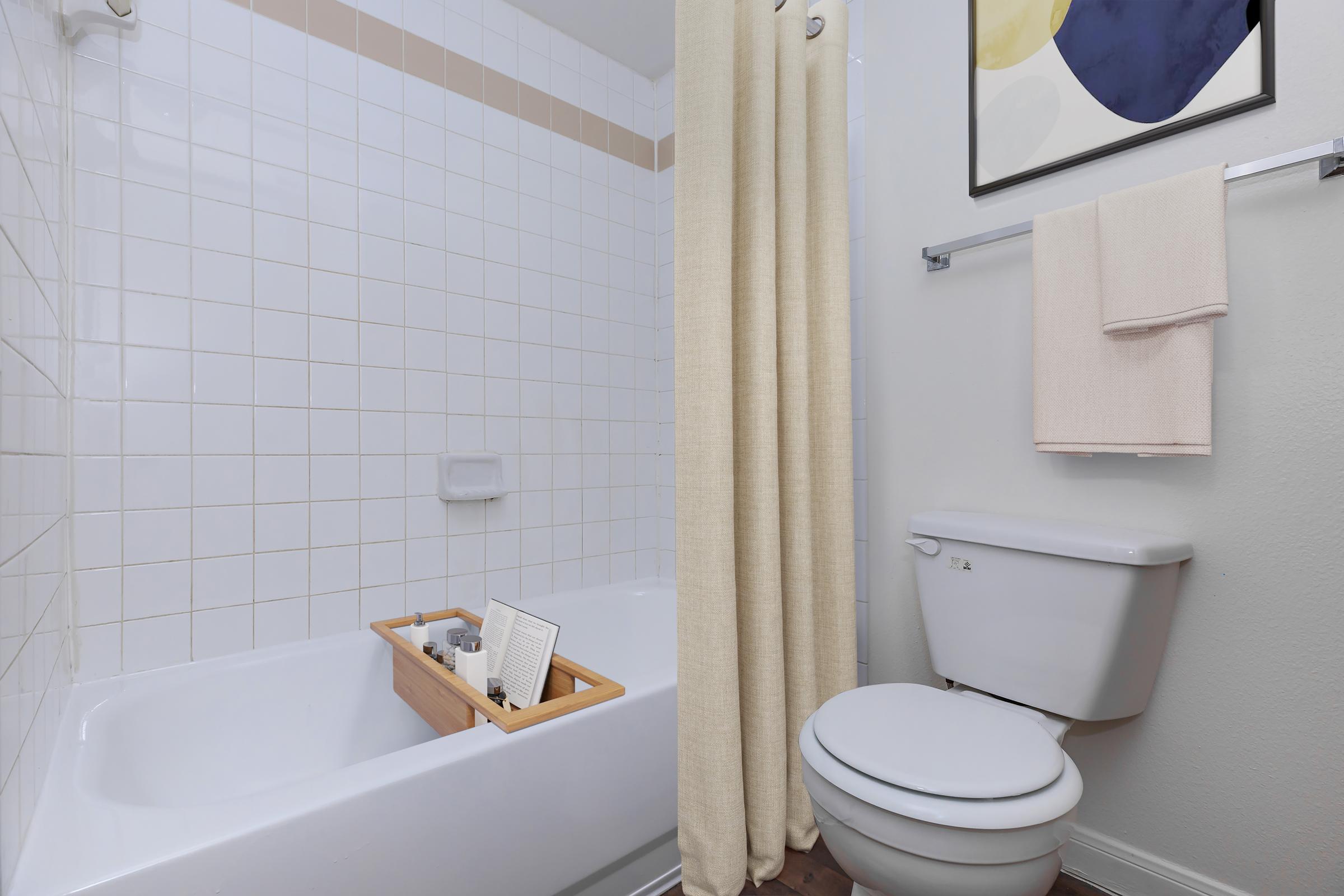 A clean and modern bathroom featuring a bathtub with a wooden tray holding a book and toiletries. A shower curtain is partially drawn. To the right, there is a white toilet and a towel rack with a neatly folded towel. A framed artwork is hung on the wall above the tub.
