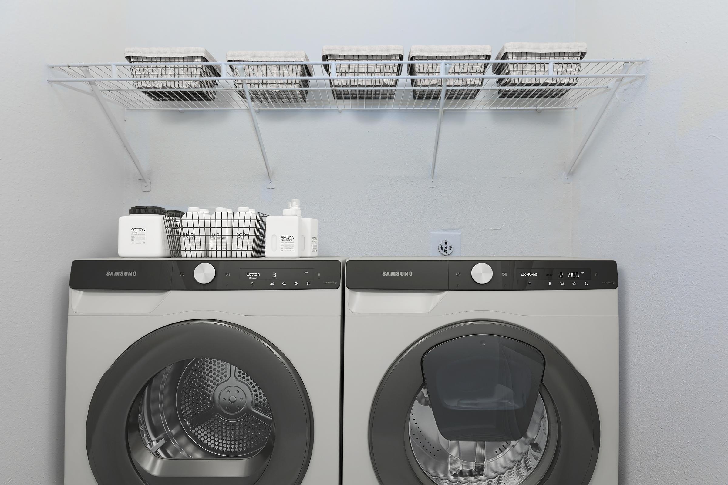 A clean laundry room featuring a pair of modern Samsung washer and dryer machines. Above them is a wall-mounted wire shelf holding several storage baskets and laundry supplies, creating an organized and functional space. The walls are painted light and the overall atmosphere is bright and tidy.