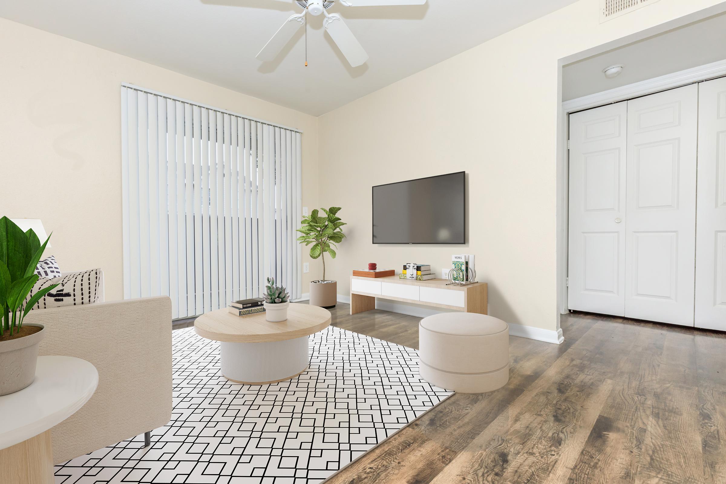 Modern living room featuring a light-colored wall and hardwood floors. A comfortable sofa with patterned cushions, a round coffee table, and a decorative plant are present. A flat-screen TV is mounted on the wall above a simple media console. Light filters through vertical blinds, and a closet door is visible in the background.
