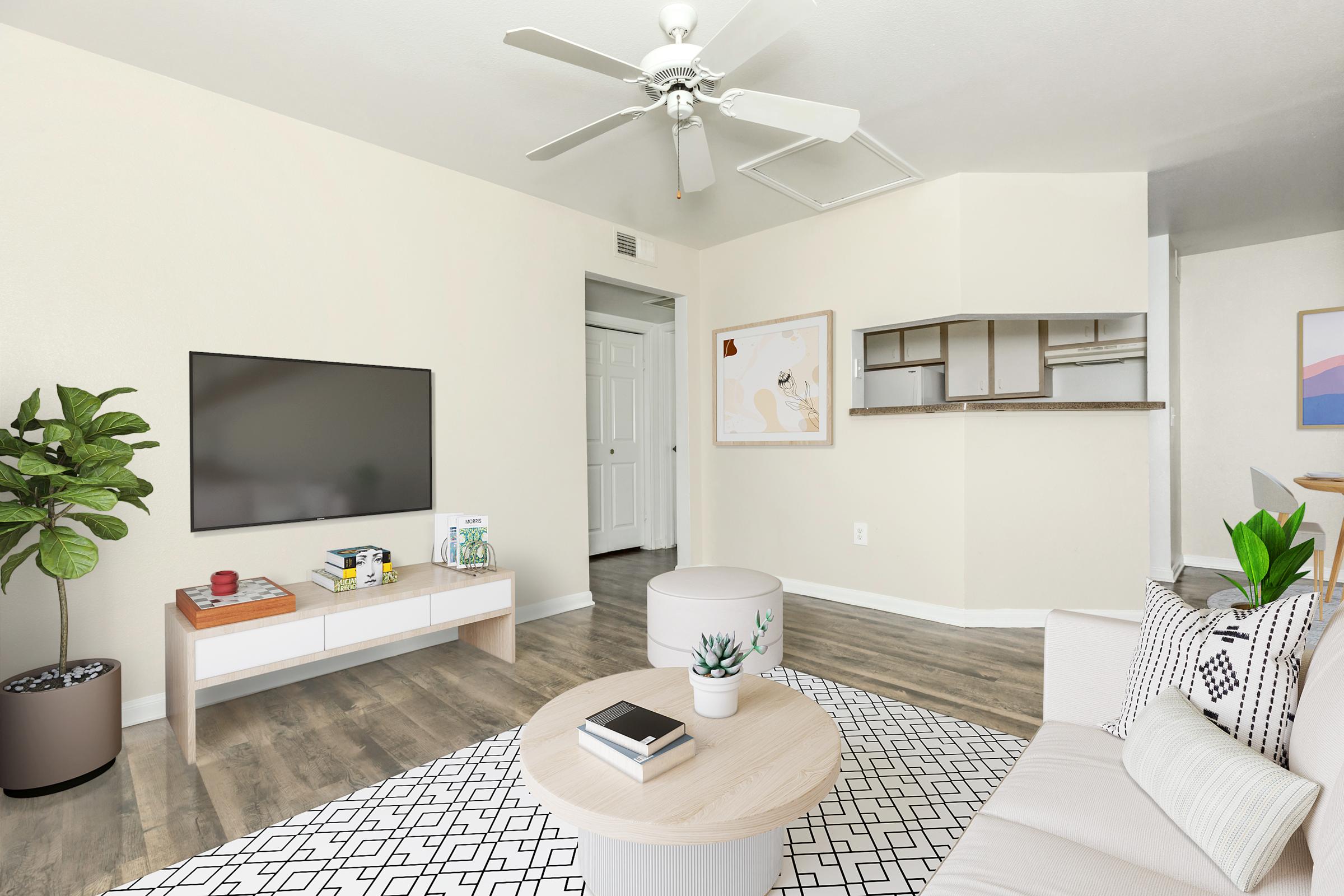 A modern living room featuring a beige couch, a round coffee table, and a large TV mounted on the wall. There's a plant in a pot next to the TV stand, a patterned area rug on the floor, and art on the wall. A doorway leads to another room, and the overall decor is light and inviting.