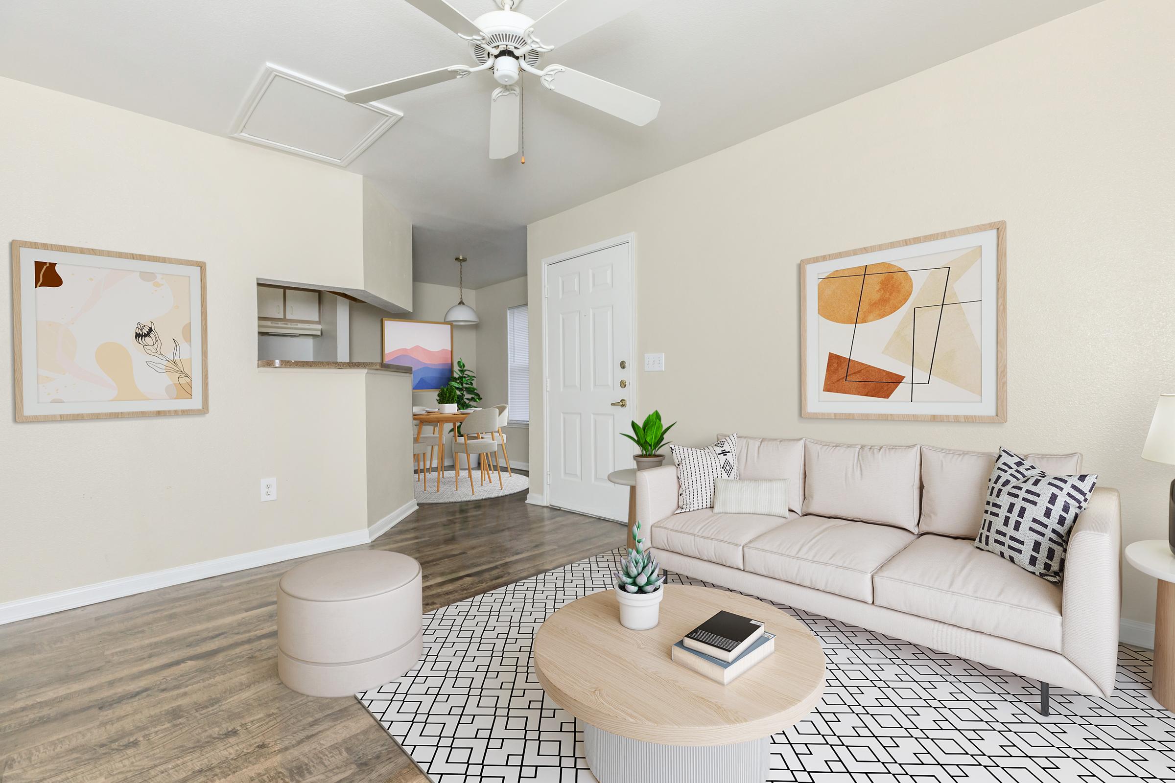 A modern living room featuring a light-colored sofa with patterned cushions, a round ottoman, and a coffee table. Artwork adorns the walls, while a fan hangs from the ceiling. A dining area is visible in the background, including a small table and chairs, with a plant adding a touch of greenery. The space has a bright and inviting atmosphere.