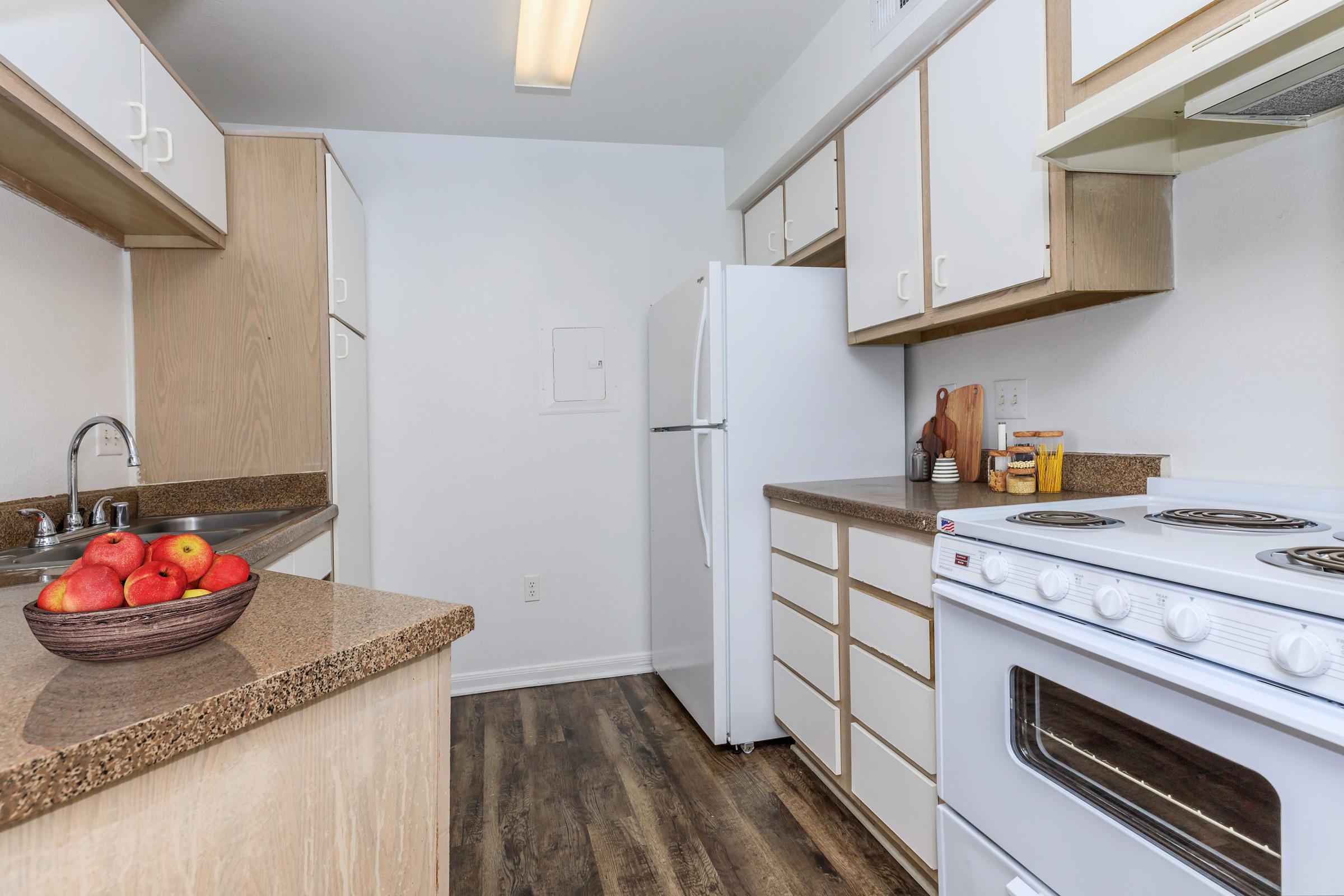 A modern kitchen with light-colored cabinets and a brown countertop. The kitchen features a white refrigerator, a white stove, and a bowl of red apples on the counter. The floor is dark wood, and there is ample natural light in the space.