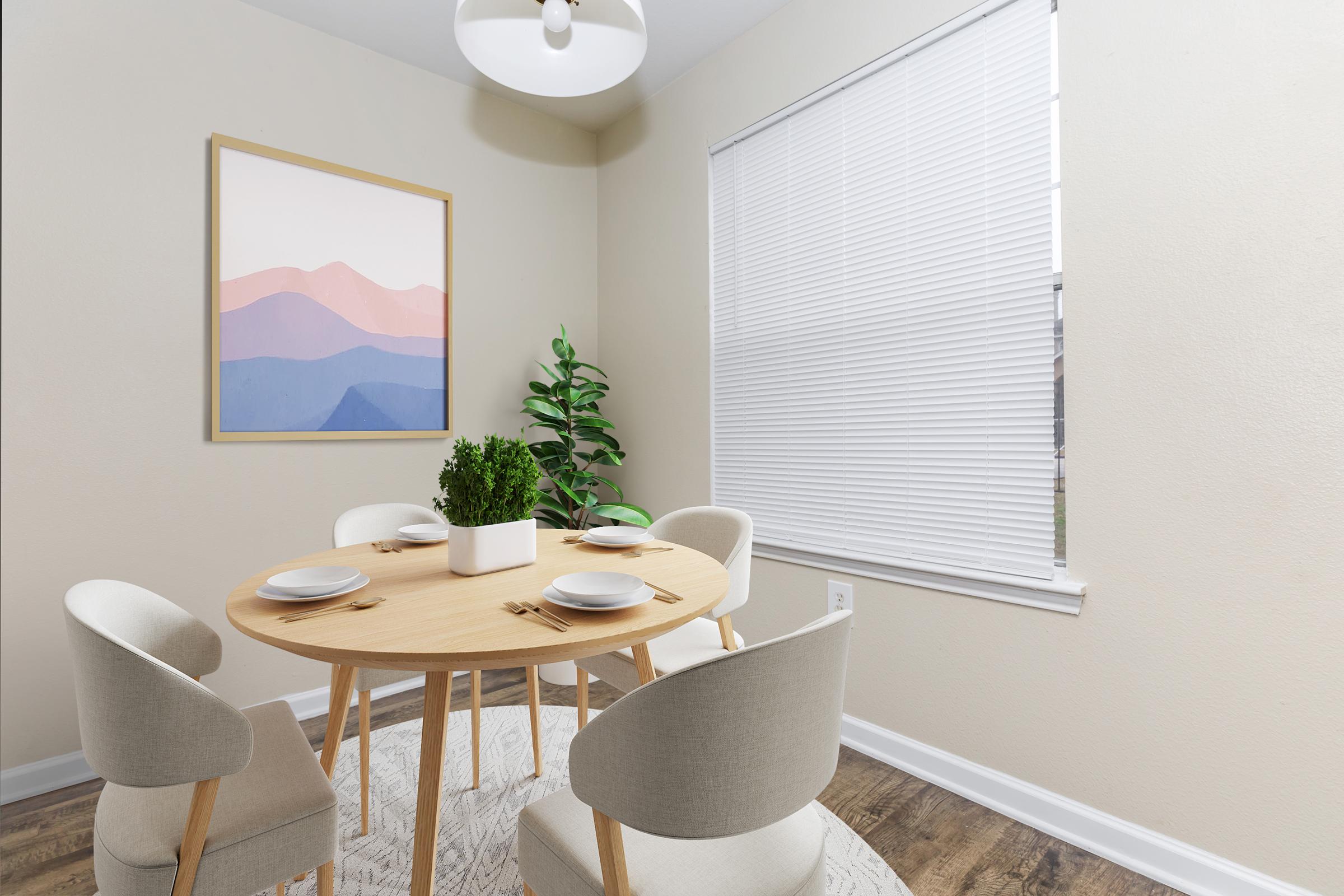 A cozy dining area featuring a round wooden table set for four with plates and cutlery. Soft beige walls complement a large framed artwork of pastel mountains. A potted plant adds greenery, while a window with blinds lets in natural light. A subtle rug completes the inviting atmosphere.