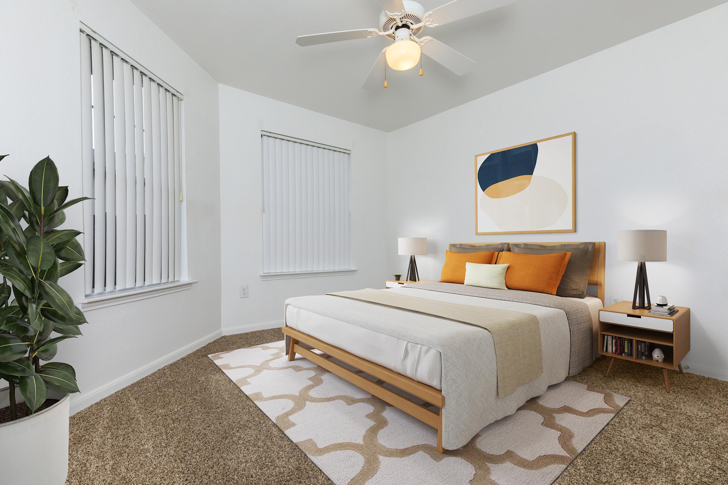 A cozy bedroom featuring a queen-sized bed with a light-colored comforter and orange accent pillows. There are two bedside lamps on wooden nightstands. A large piece of abstract art hangs on the wall. The room has light-colored walls, a ceiling fan, and a potted plant in the corner, along with a patterned rug on the floor.