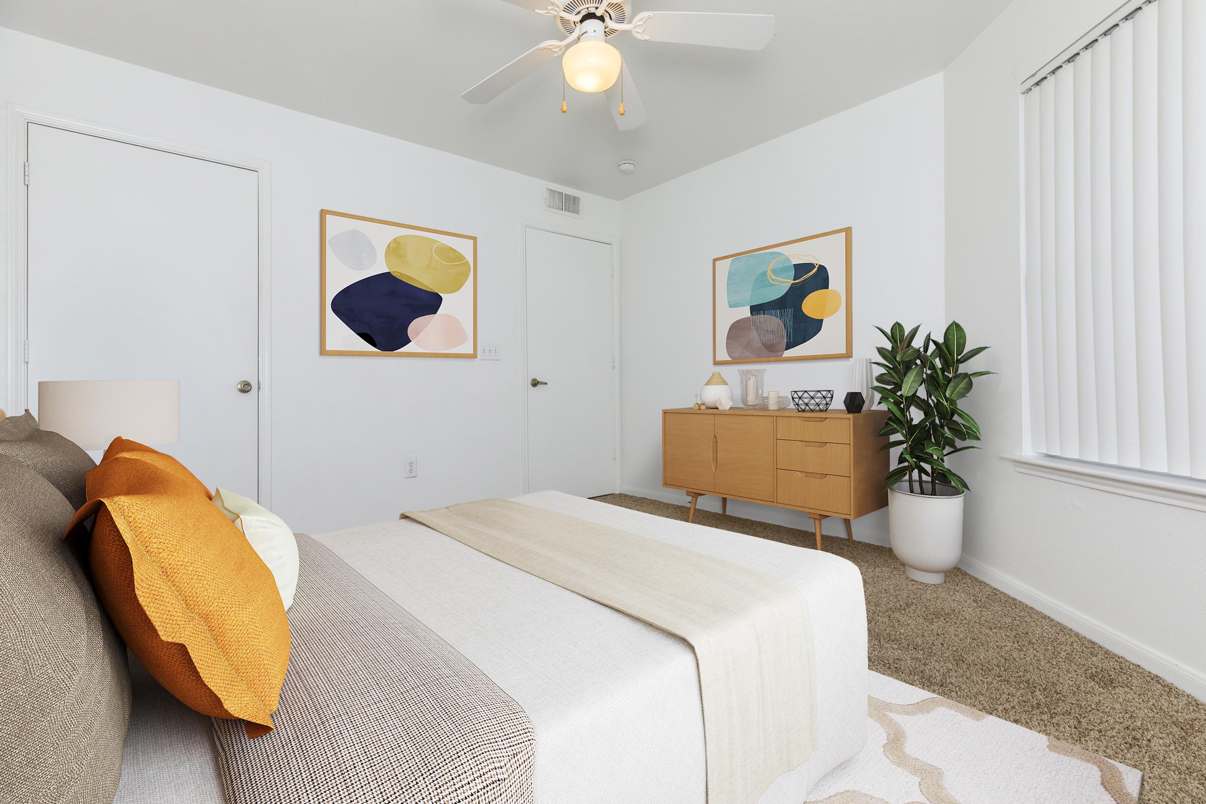 A stylish and modern bedroom featuring a bed with decorative pillows, a light-colored throw blanket, and a wooden dresser. Two framed abstract art pieces hang on the wall, and a bright green plant is positioned near the dresser. The room has neutral colors and natural light from a window with blinds.