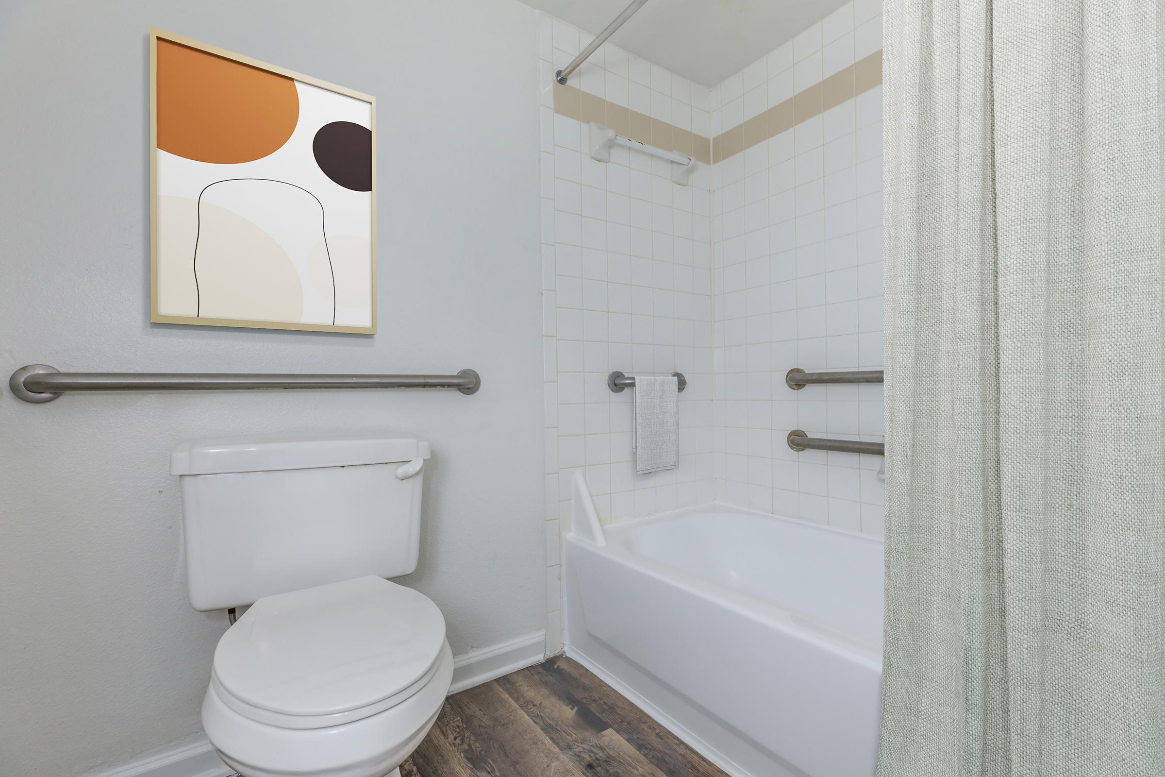 A clean bathroom featuring a white toilet, a bathtub with a shower curtain, and grab bars for safety. A minimalistic abstract art piece hangs on the wall, adding a splash of color with warm tones. The floor has a wooden appearance, complementing the light-colored walls and tiles.