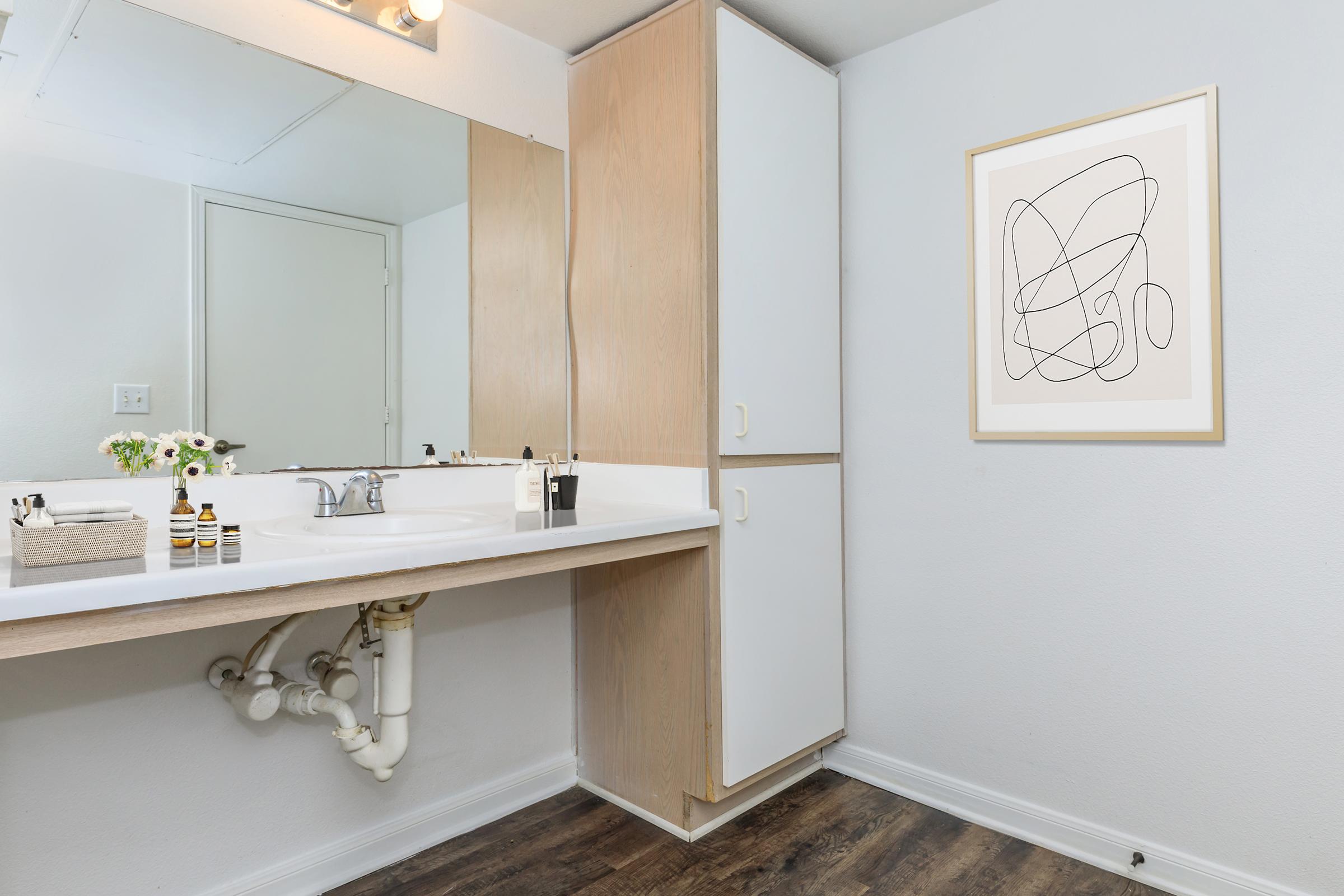 A clean and modern bathroom featuring a white sink with a wooden countertop, a large mirror above, and minimalistic decor. There are two cabinets, one of which is closed. A framed abstract artwork hangs on the wall, and a small arrangement of flowers adds a touch of freshness to the space.