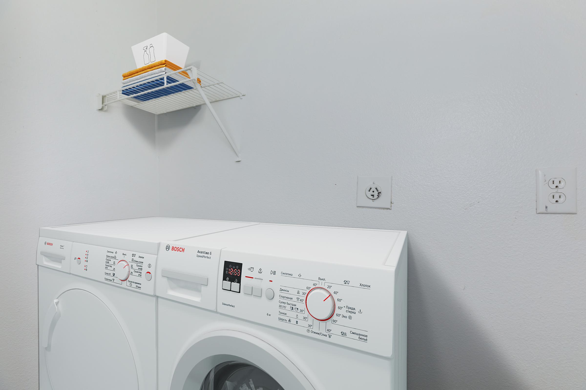 A laundry room featuring a white Bosch washer and dryer side by side, with control dials and displays visible. Above them, a wire shelf holds a few items, including a white container and a small stack of yellow bins. The wall is painted a light color, and electrical outlets are present.