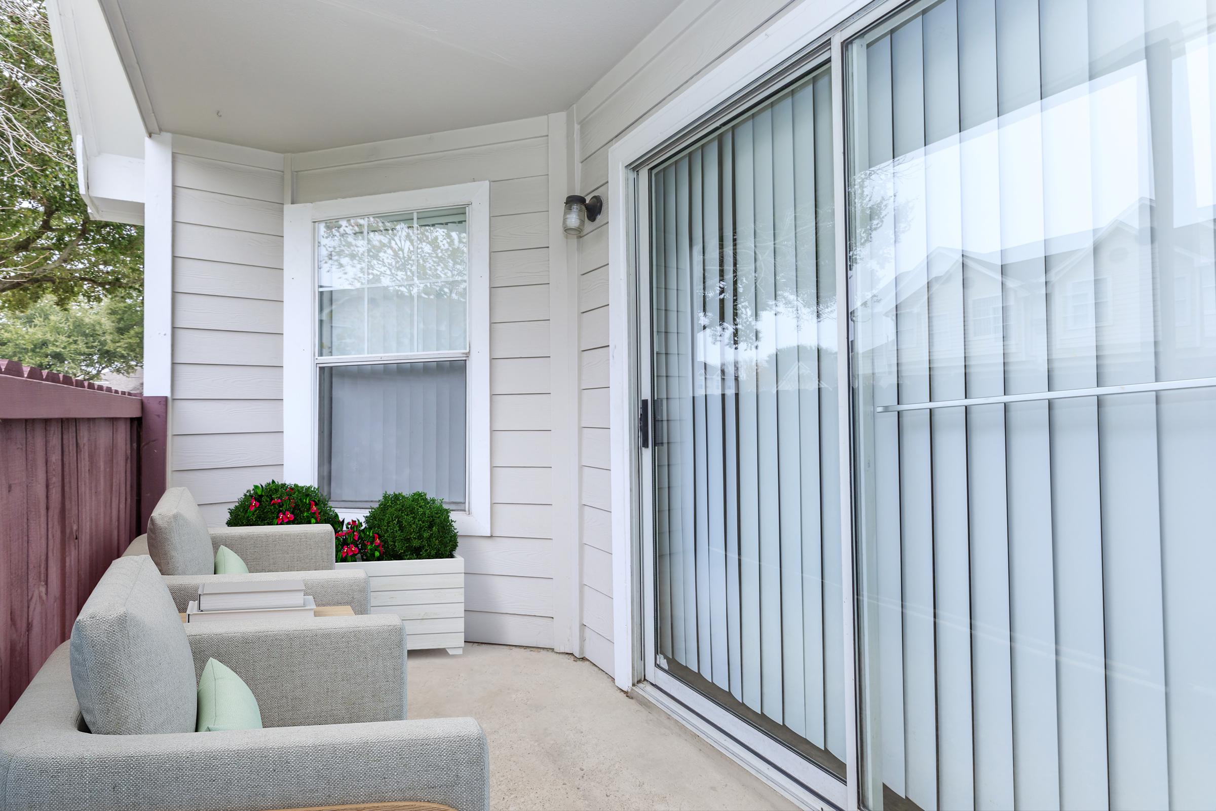 A small outdoor patio area with two light gray chairs and potted plants. The patio features sliding glass doors with vertical blinds, allowing natural light to enter. Fenced with a wooden structure, it provides a cozy and private atmosphere for relaxation.