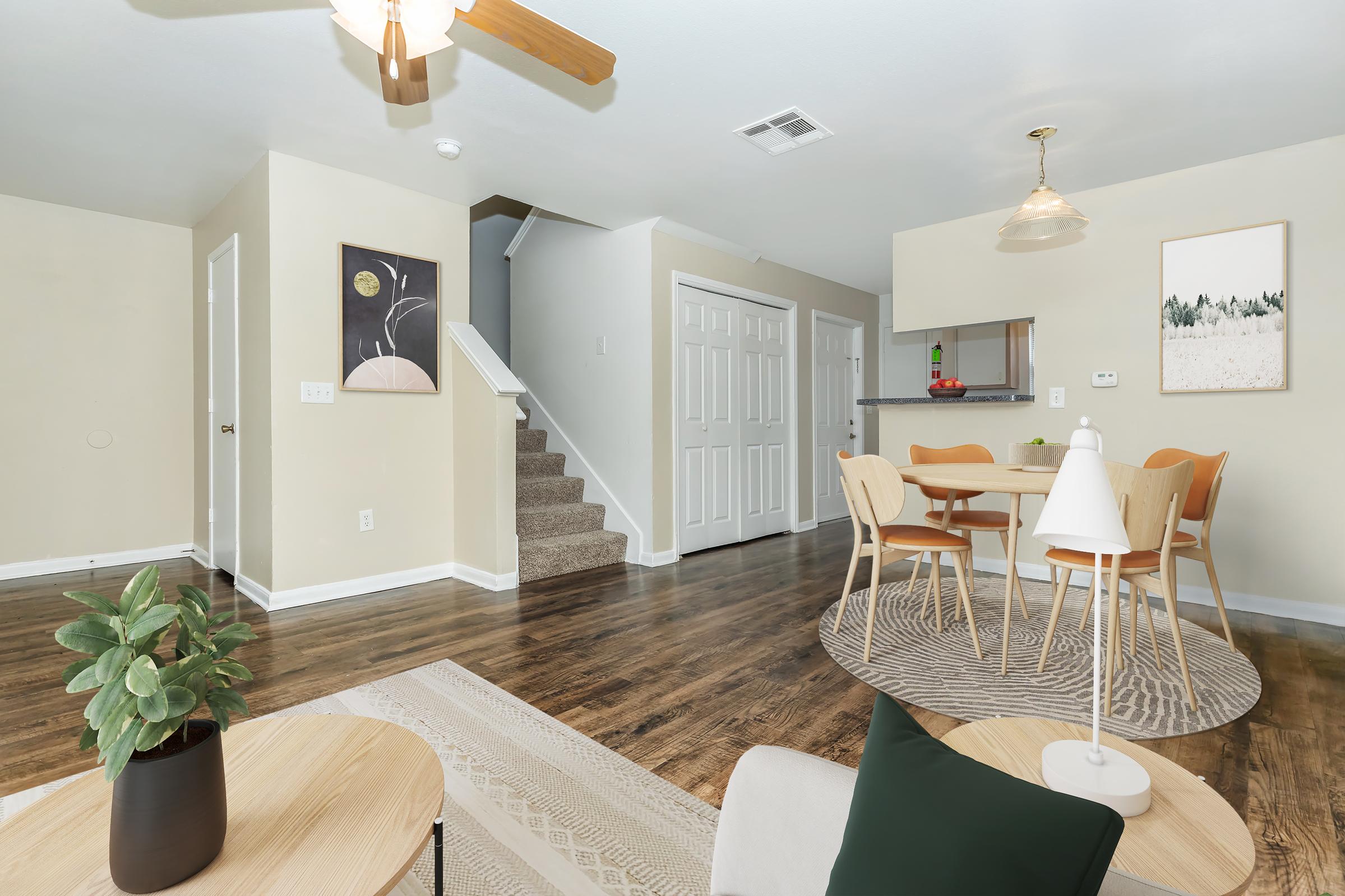 A well-decorated living space featuring a light-colored wall, a wooden floor, and a staircase. A dining area with a round table and chairs is visible, accompanied by modern decor elements like a plant and artwork. Soft lighting from a ceiling fan and a pendant light creates a warm ambiance.