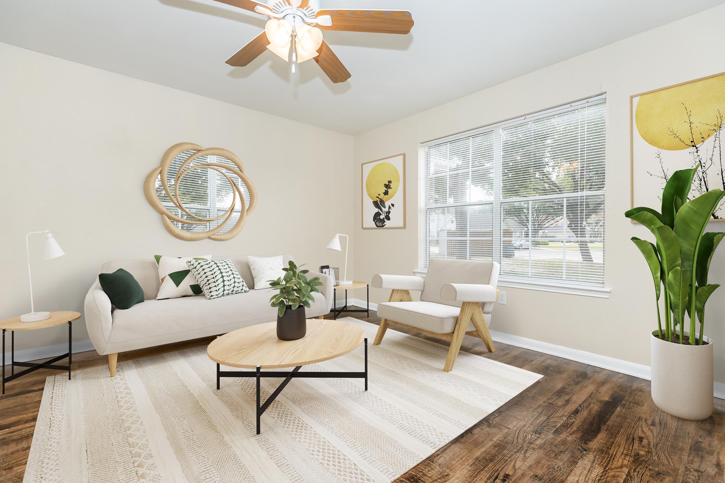 Stylish living room featuring a beige couch with green and white pillows, a round coffee table, and a large mirror. A plant is positioned in the corner beside a bright window with natural light. Artwork with yellow and green accents adorns the walls, and a ceiling fan adds a cozy touch to the space.