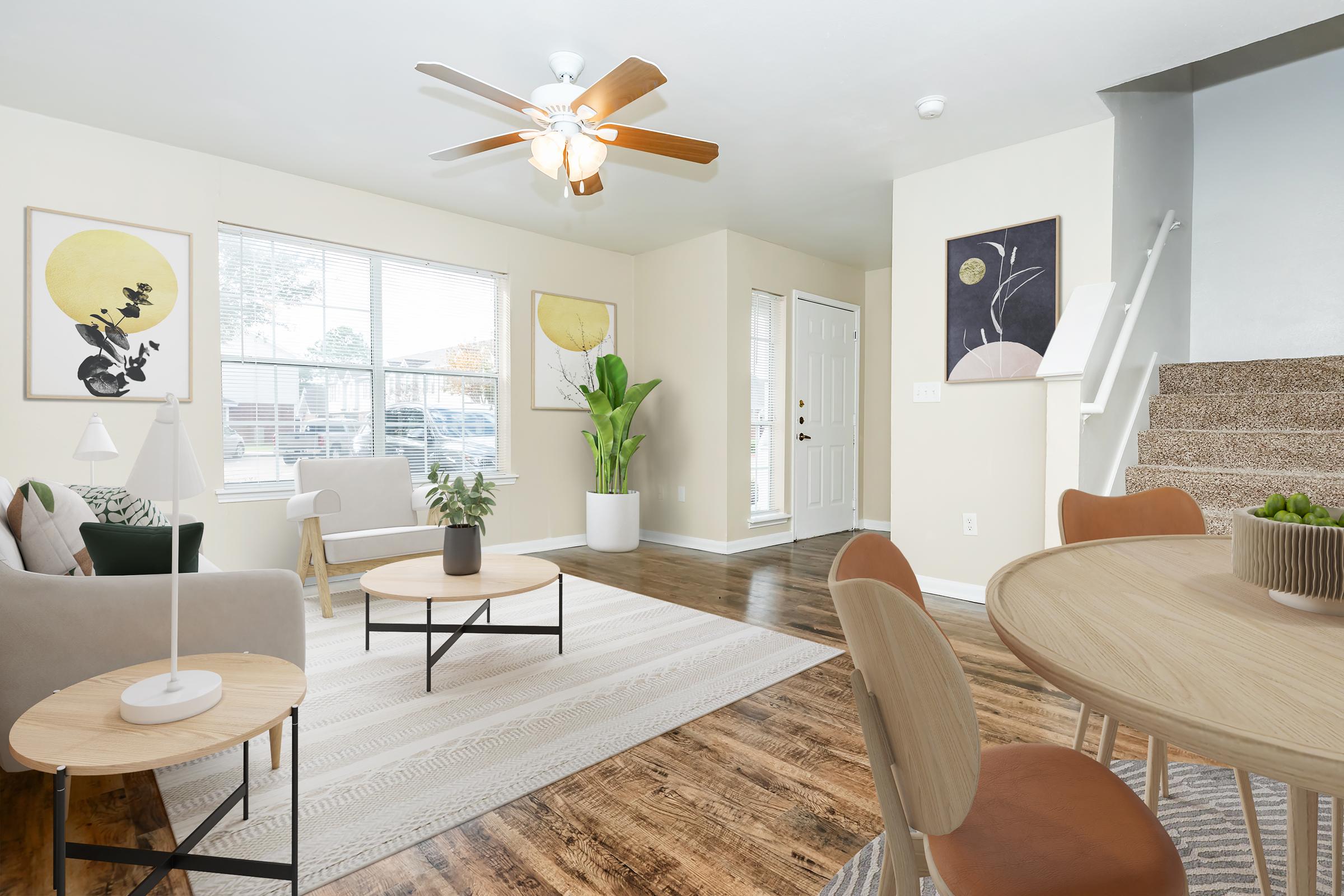 Bright living room featuring a beige sofa, wooden coffee tables, and a round table with chairs. Decor includes art with circular designs on the walls, a potted plant, and a ceiling fan. The space is airy with natural light from large windows and a welcoming entrance.