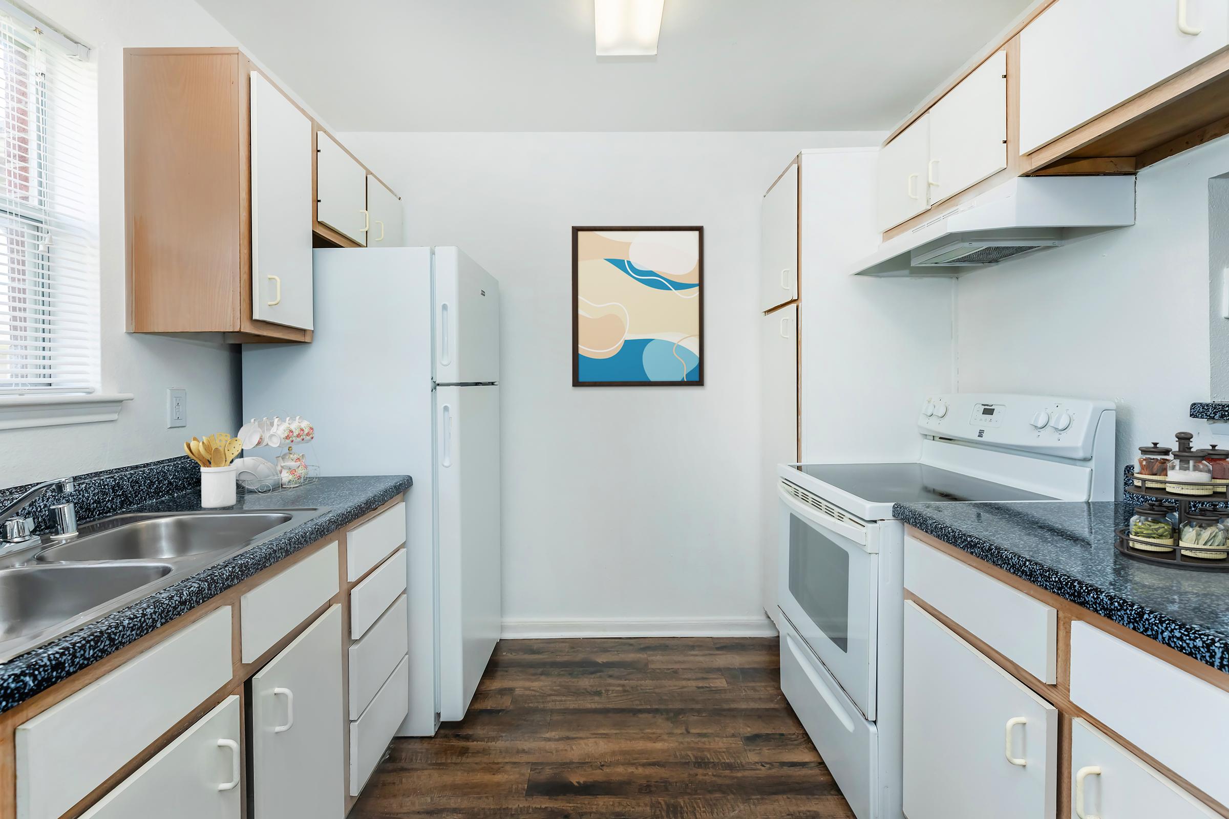 A modern kitchen featuring white cabinetry, a double sink, a refrigerator, and an oven. The kitchen has dark countertops and wooden flooring. A framed abstract art piece hangs on the wall, adding a touch of color to the space. Natural light comes in from a window, creating a bright atmosphere.