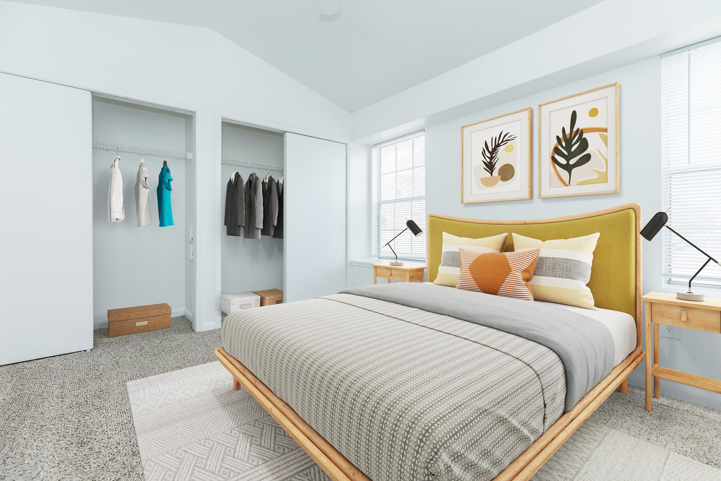 A well-decorated bedroom featuring a yellow bed frame with gray and orange bedding. There are two bedside tables with lamps, and behind the bed, there are framed wall art pieces. A closet is visible on the left with neatly hung clothes, and natural light enters through the windows, enhancing the cozy atmosphere.