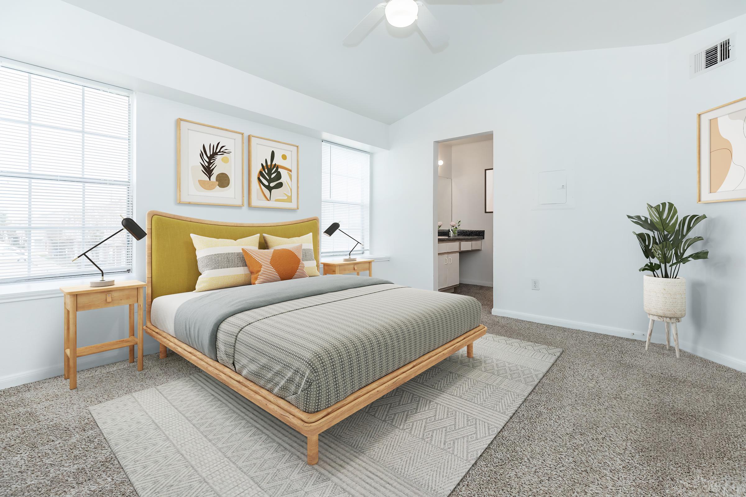 A bright and modern bedroom featuring a large bed with a yellow headboard, grey and white bedding, and decorative pillows. There are two wooden nightstands with lamps on either side. The room has large windows allowing natural light, and a potted plant adds a touch of greenery. The walls are painted white, and there's a rug under the bed.