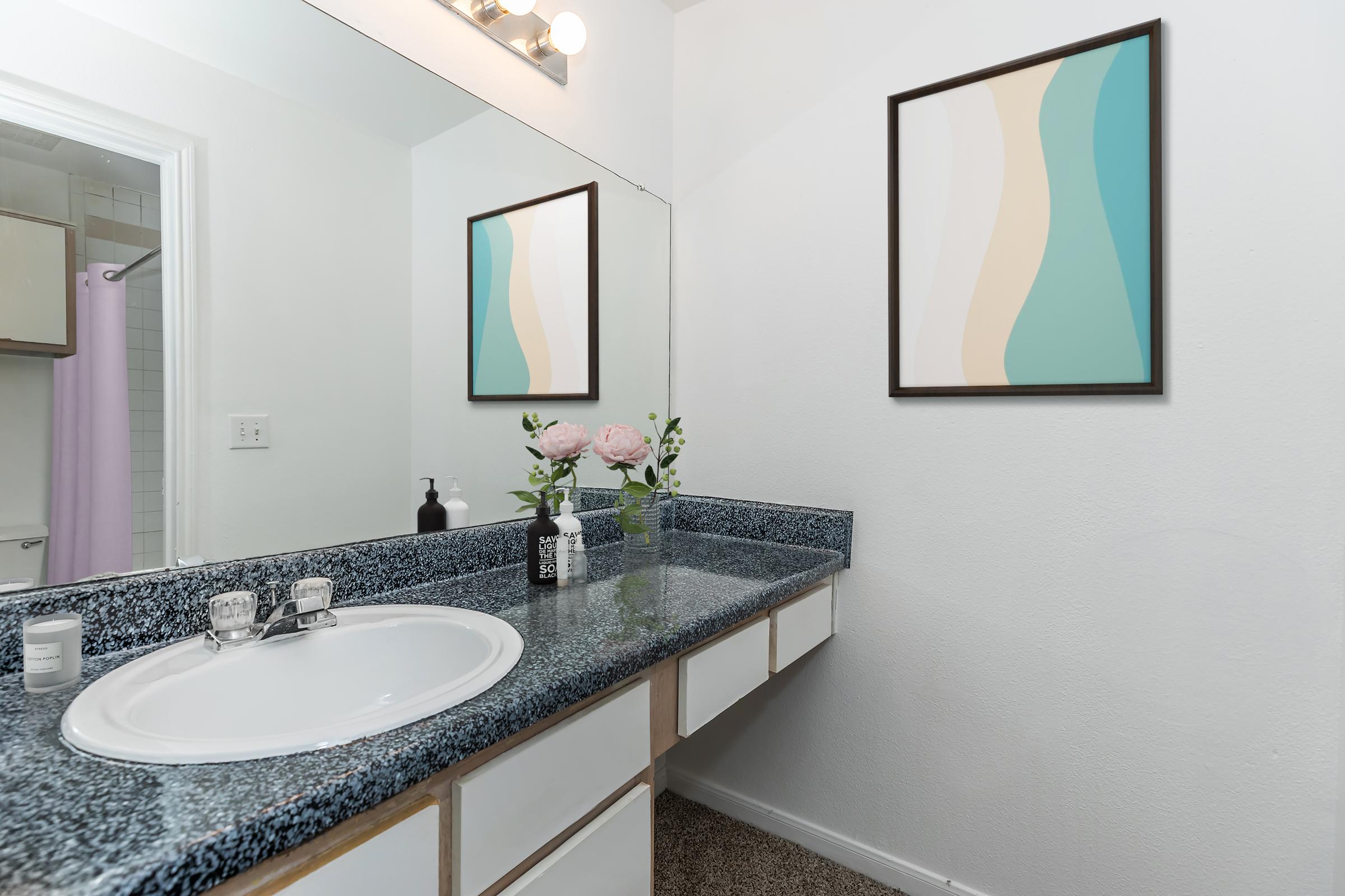 A modern bathroom featuring a blue speckled countertop with a white sink, framed artwork on the wall, and a vase of pink peonies. The space has light-colored walls and a mirror above the sink, with a shower curtain visible in the background.