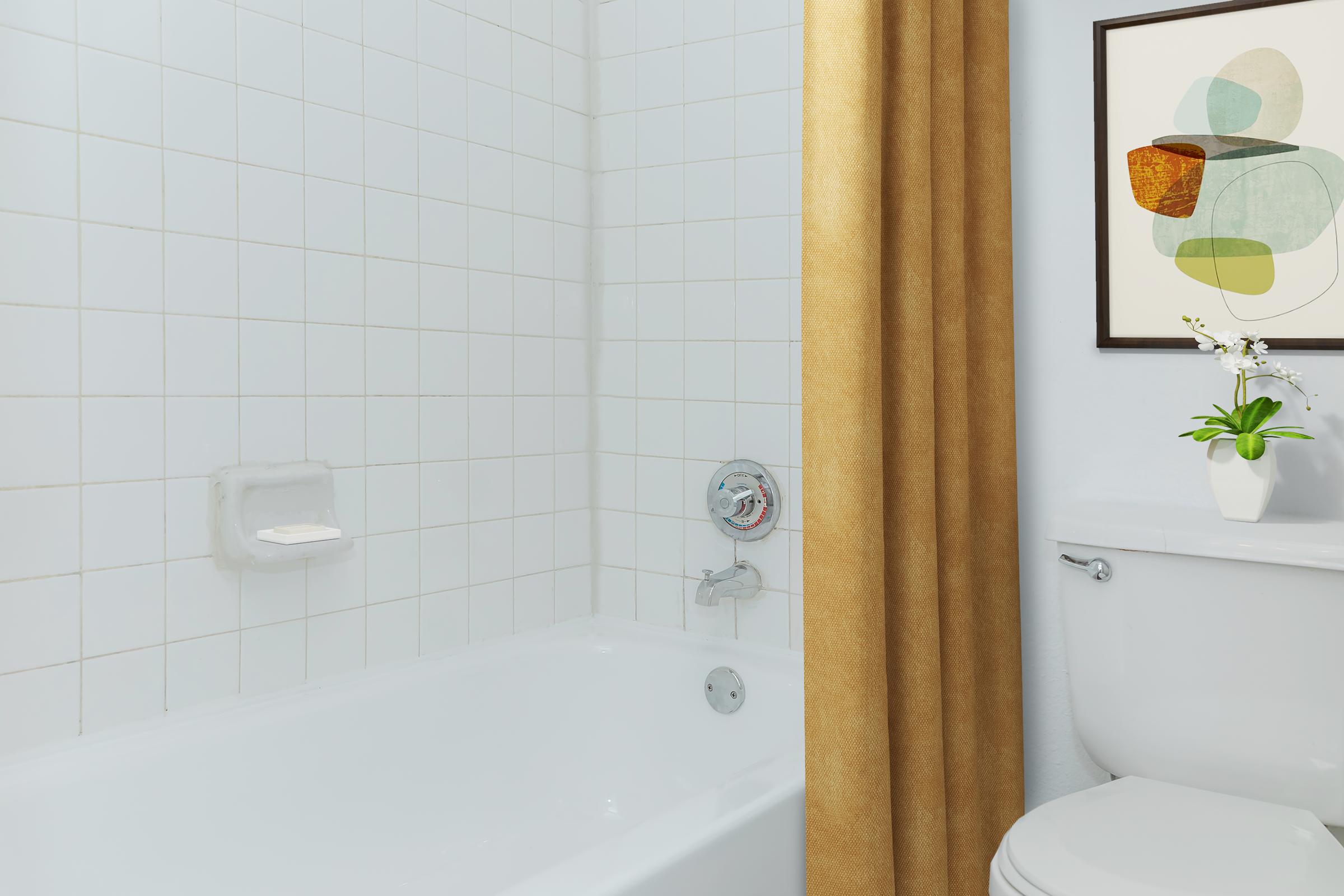 A clean bathroom featuring a white tiled bathtub with a beige shower curtain, a white toilet, and a small potted plant on a bathroom countertop. A piece of abstract artwork hangs on the wall above the bathtub, adding a touch of color to the serene space.