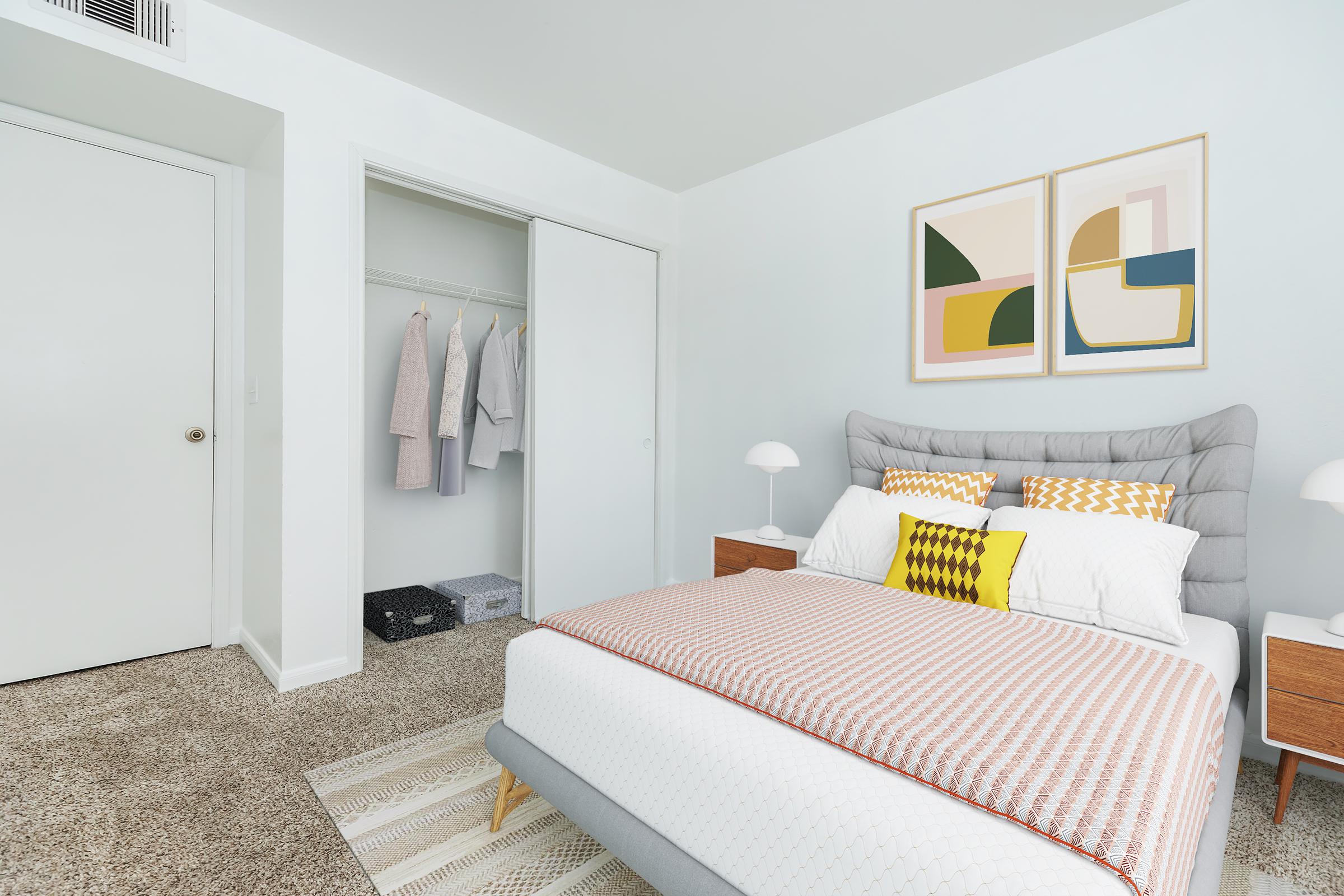 A bright and modern bedroom featuring a gray upholstered bed with colorful pillows, a stylish patterned throw, and nightstands on either side. A closet with hangers is visible, and abstract artwork adorns the wall. The room has a light color scheme and carpeted flooring, creating a cozy atmosphere.