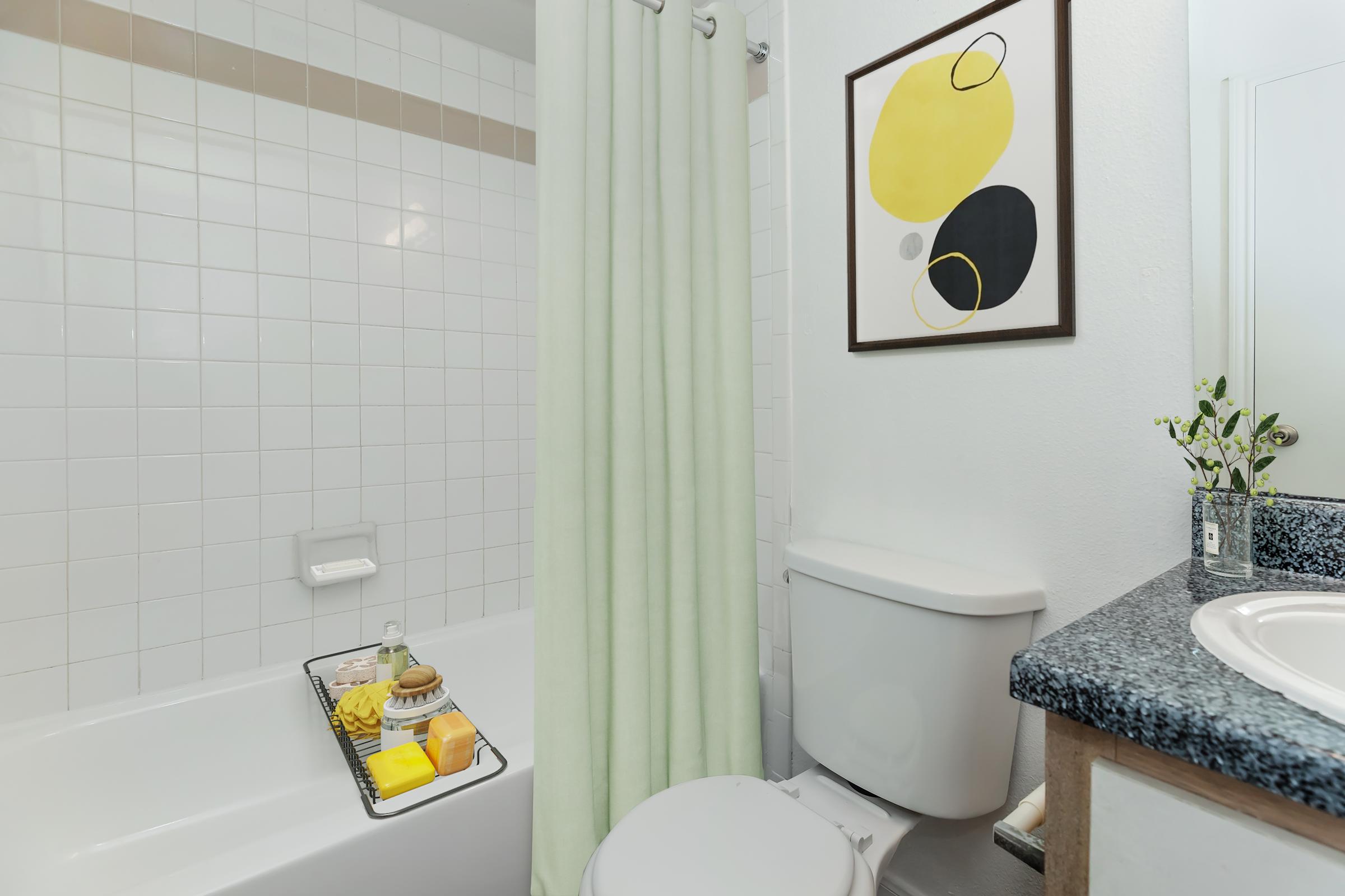 A clean, modern bathroom featuring a bathtub with a shower curtain, a toilet, and a vanity. On the bathtub, there's a tray with bath essentials like soap and candles. A piece of abstract wall art with yellow and black shapes hangs above the toilet, adding a pop of color to the space.