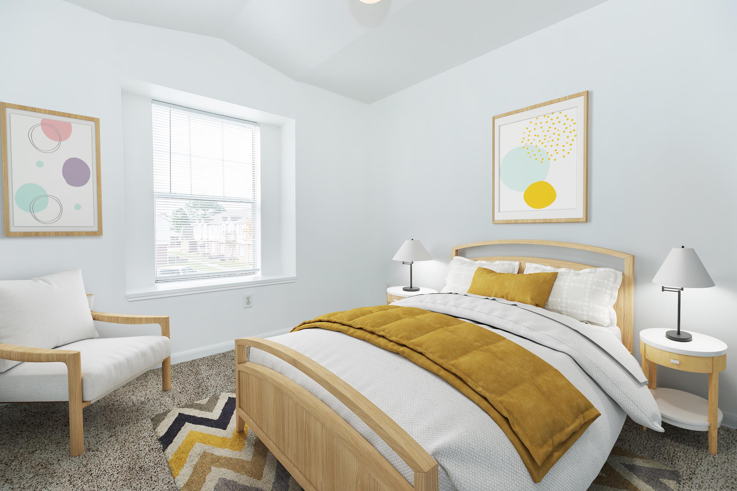 A cozy bedroom featuring a comfortable bed with a yellow throw blanket, bedside tables with lamps, and a chair in the corner. The walls are painted white, and there are two framed abstract artworks above the bed. A window lets in natural light, offering a view of the outside. The floor is covered with carpet and a patterned area rug.