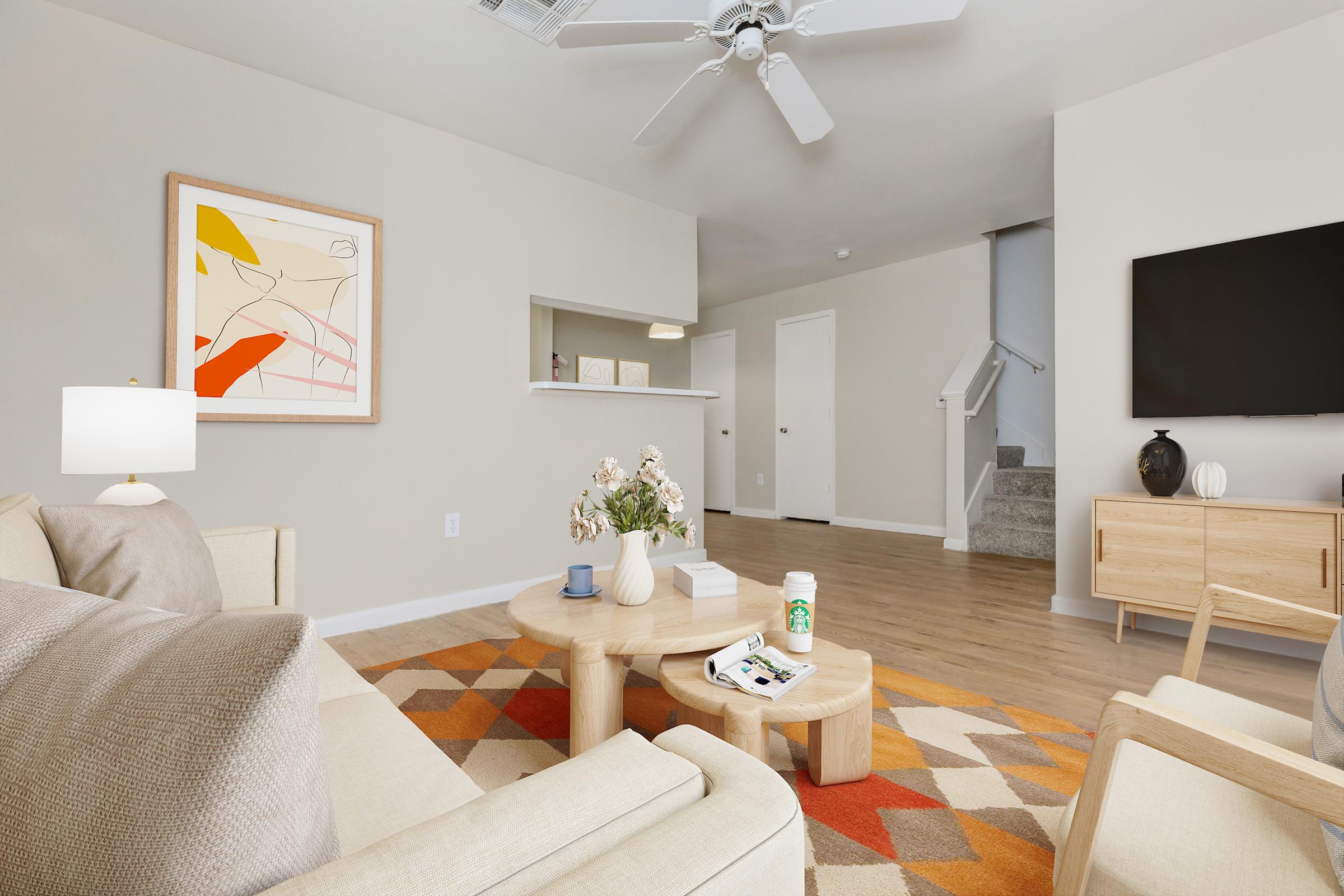 A modern living room featuring a light-colored sofa and a wooden coffee table. There are decorative items on the table, including a magazine and a drink. The room has a colorful area rug, a wall-mounted TV, and art on the wall. A ceiling fan is visible, along with a staircase in the background.