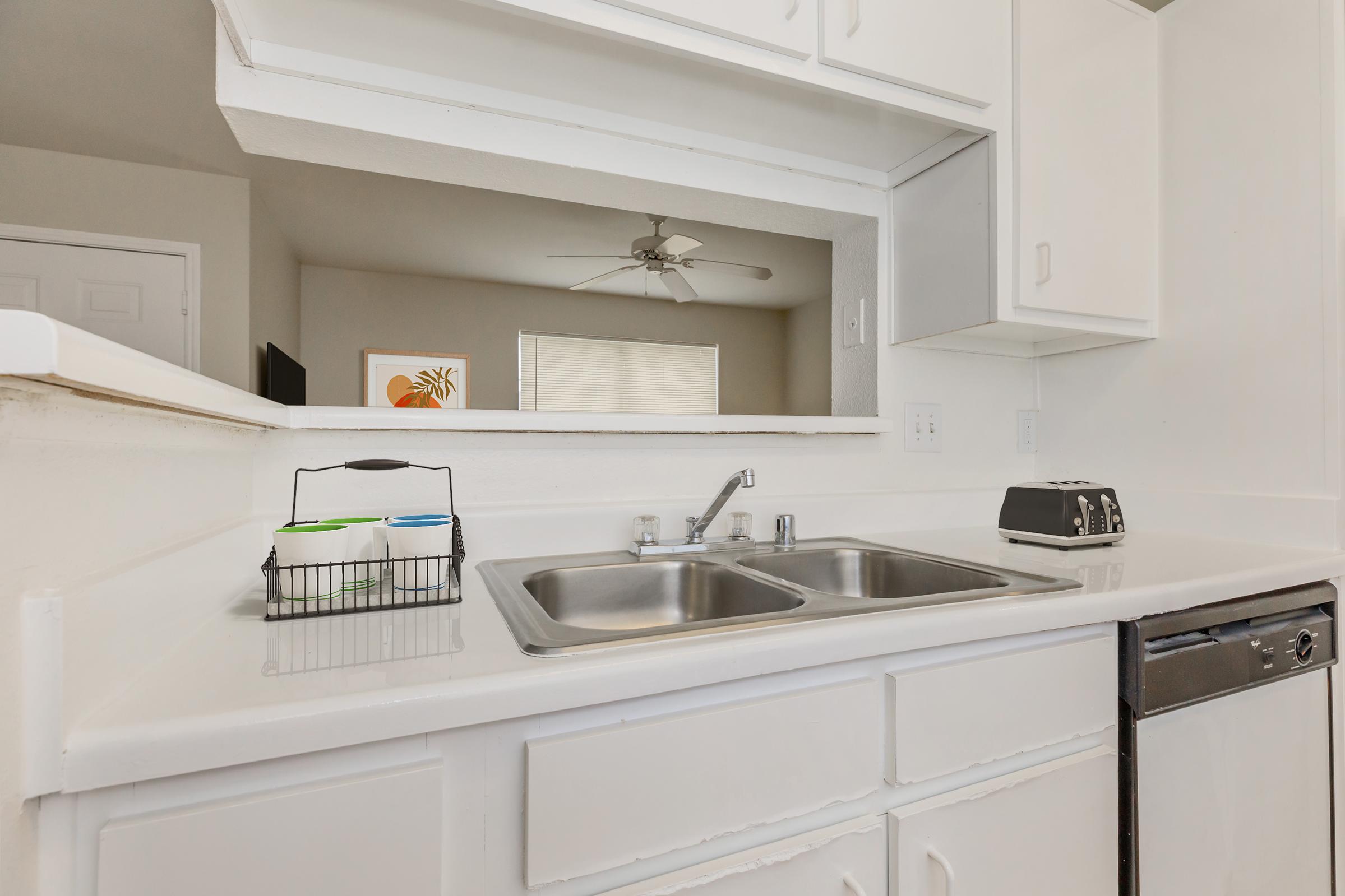 A clean kitchen countertop with a double sink, a black toaster, and a wire basket containing colorful dishware. A window is visible in the background, along with a ceiling fan, creating a bright and airy atmosphere. The cabinetry and appliances are white, contributing to a modern look.