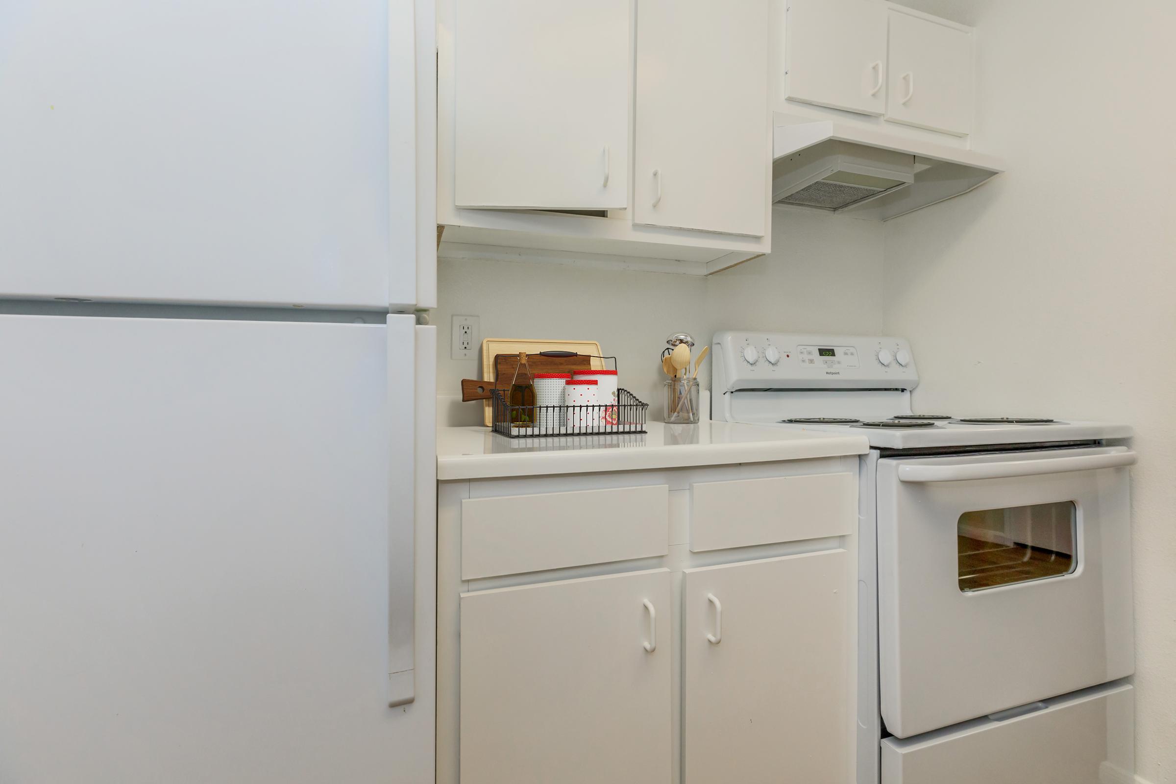 A modern kitchen featuring white cabinets, a refrigerator, an oven, and a stove. There is a countertop with a basket holding cooking utensils and various containers. The walls are painted in a light color, creating a bright and clean atmosphere.