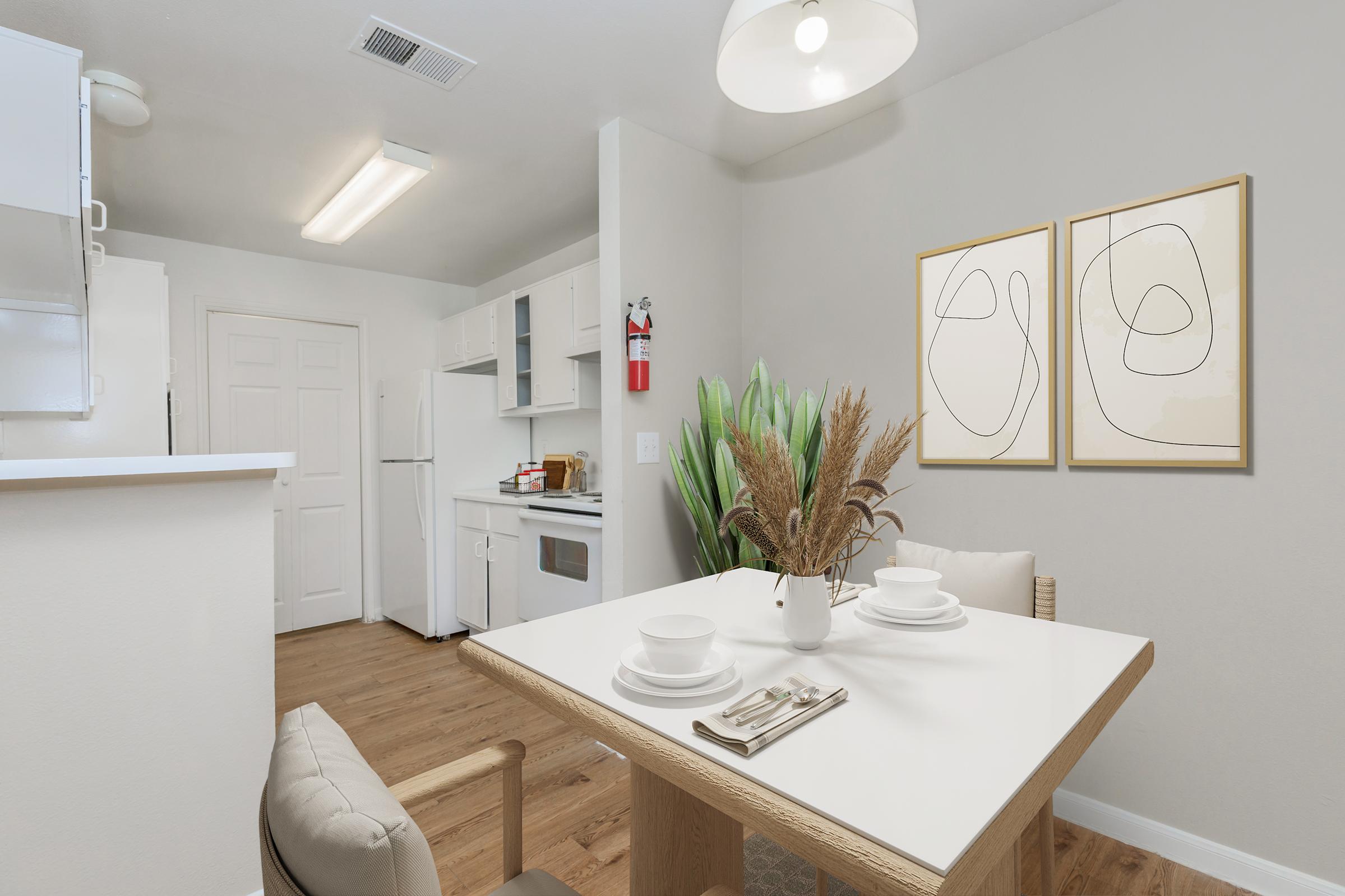 A modern kitchen and dining area featuring a white table set with plates and utensils, surrounded by comfortable chairs. The kitchen has white cabinets, a refrigerator, and a stove. Two abstract art pieces hang on the wall, and a green plant adds a touch of color to the neutral decor.