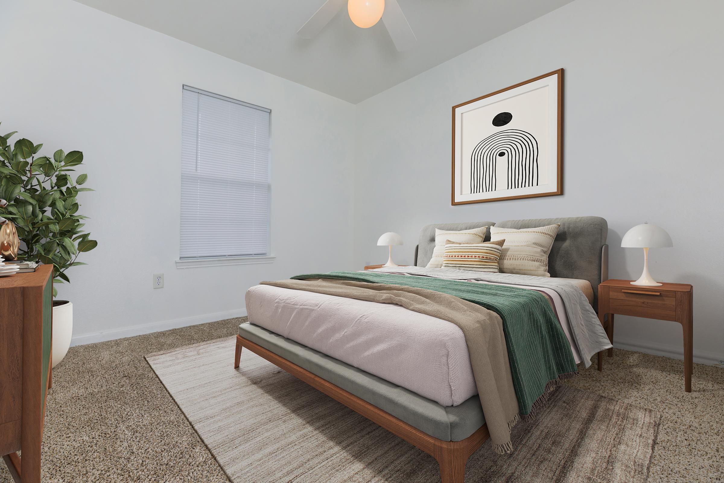 A cozy bedroom featuring a neatly made bed with multiple textured blankets and pillows. There is a stylish framed wall art above the bed, a small nightstand with a lamp on one side, and a houseplant in the corner. Natural light filters through a window, enhancing the warm, inviting atmosphere.