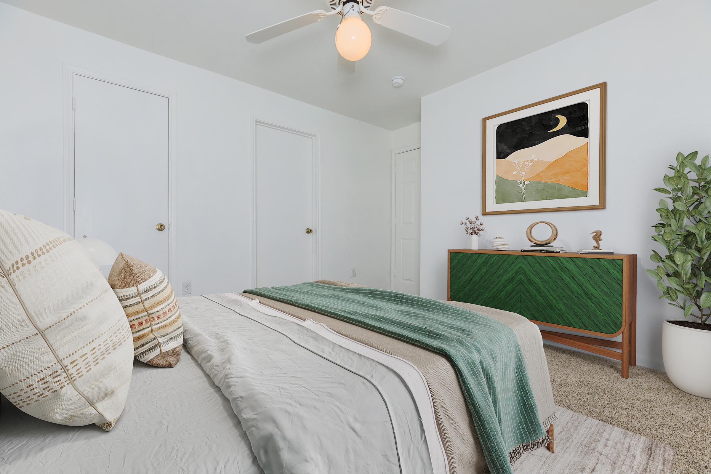 A cozy bedroom featuring a neatly made bed with decorative pillows, a soft green throw blanket, and a modern dresser. The room has light-colored walls, a ceiling fan, and a framed artwork depicting abstract landscapes. A potted plant adds a touch of greenery, while a door and closet are visible in the background.