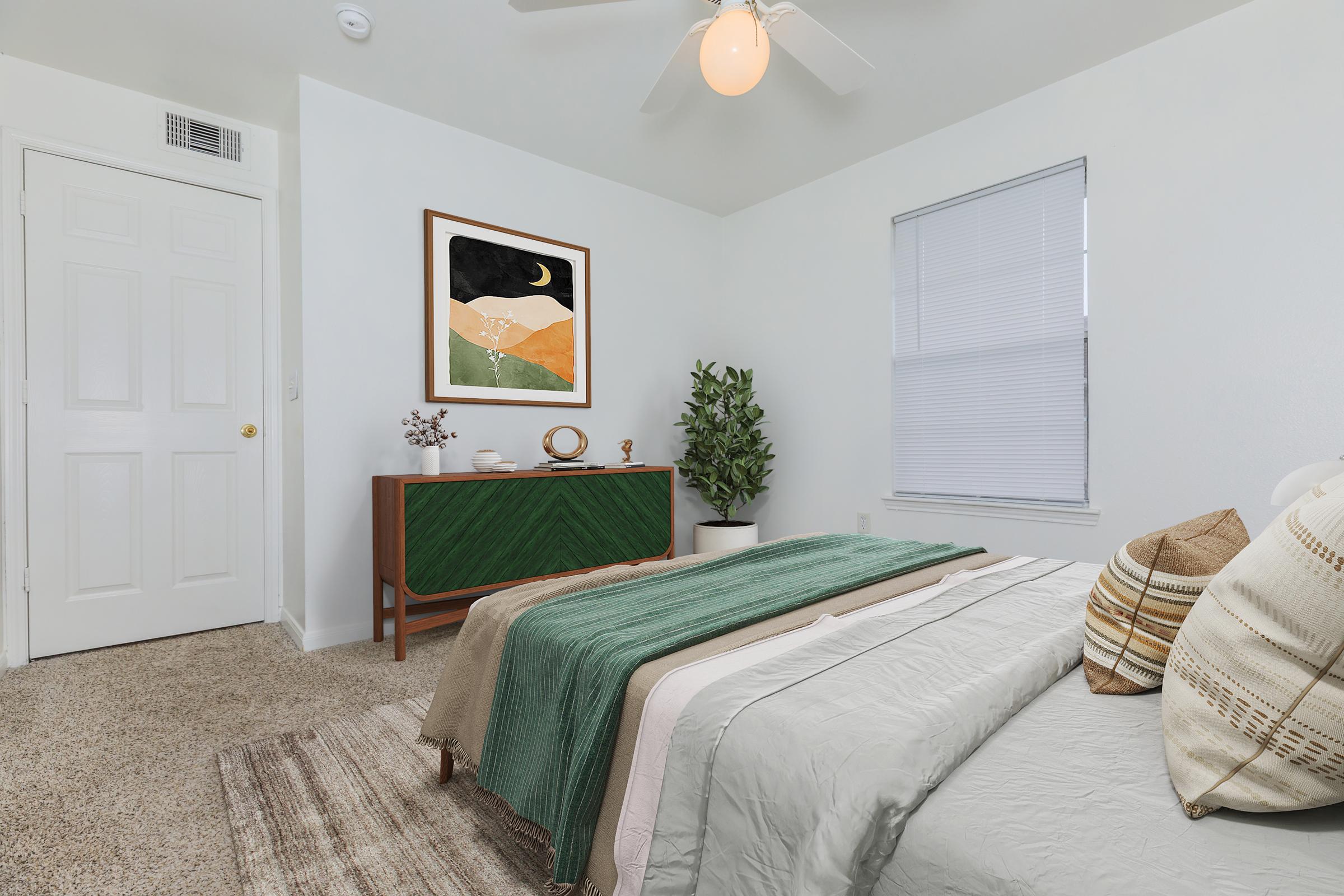 A cozy bedroom featuring a neatly made bed with textured blankets in green and beige. A stylish wooden dresser with decorative items is against the wall, and a plant adds a touch of greenery. A framed art piece hangs above the dresser, and a ceiling fan is visible. Soft natural light filters through the window.