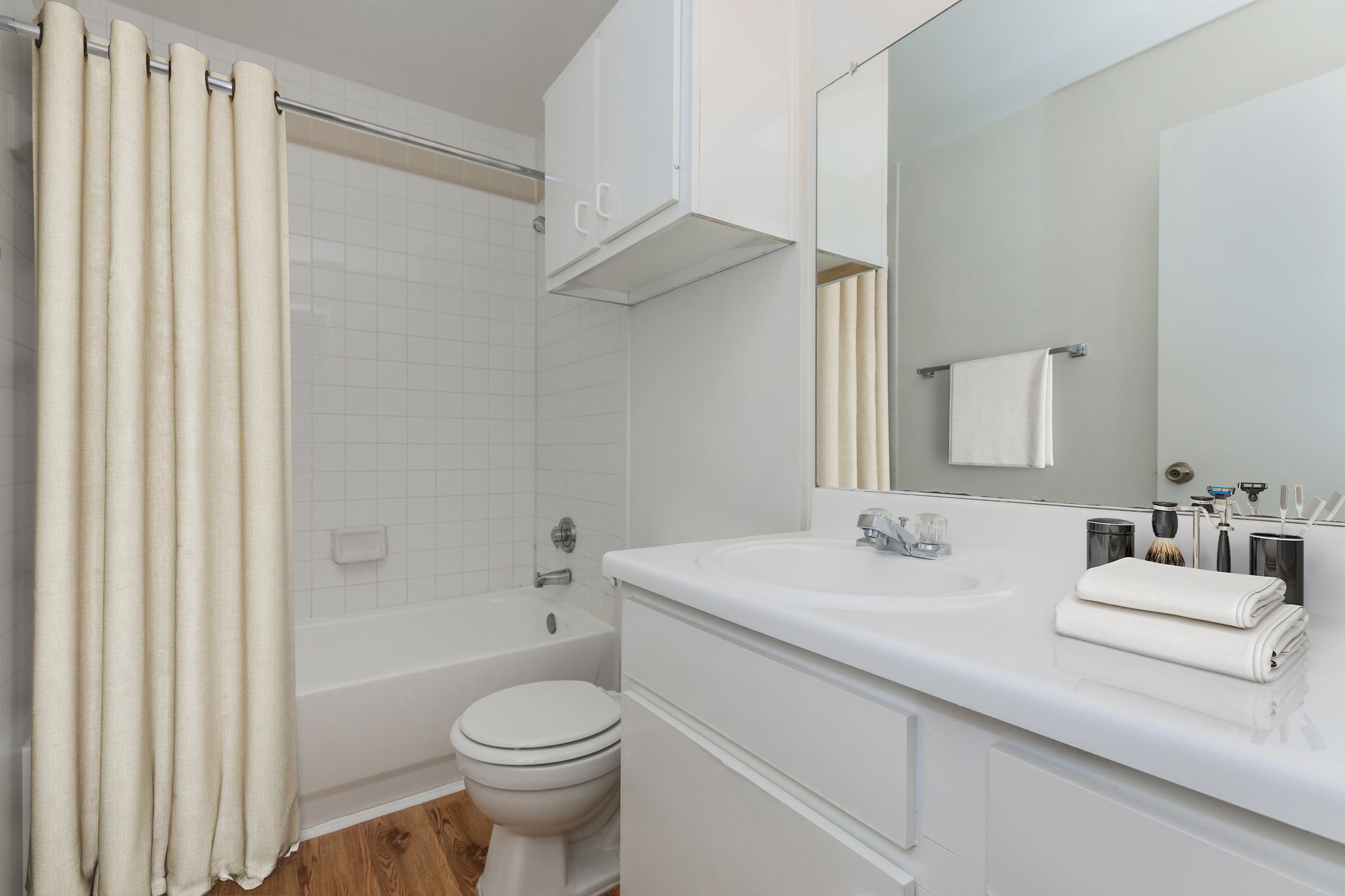 A clean and modern bathroom featuring a bathtub with a shower curtain, a white sink with a chrome faucet, and a countertop. There are neatly stacked white towels next to the sink, a towel hanging on a rack, and a large mirror above the sink. The flooring is light wood, creating a warm atmosphere.
