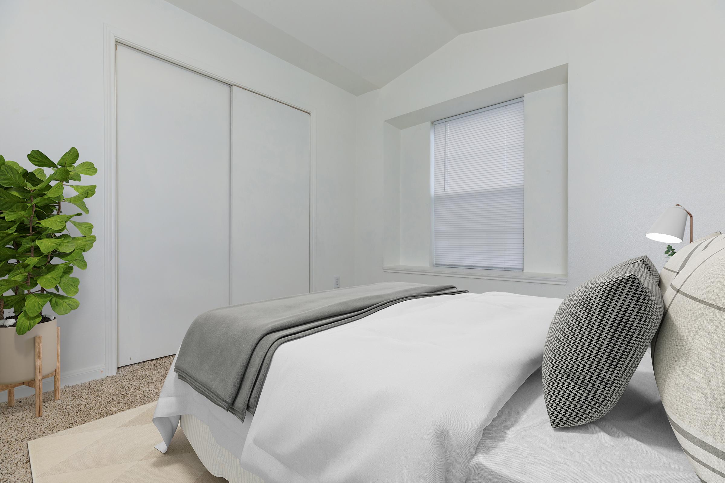 A cozy bedroom featuring a neatly made bed with gray and white bedding, a bedside lamp, and a window with blinds. A potted plant adds a touch of greenery to the space, which has light-colored walls and carpet. A closed closet is visible in the background, enhancing the room's soft and inviting atmosphere.