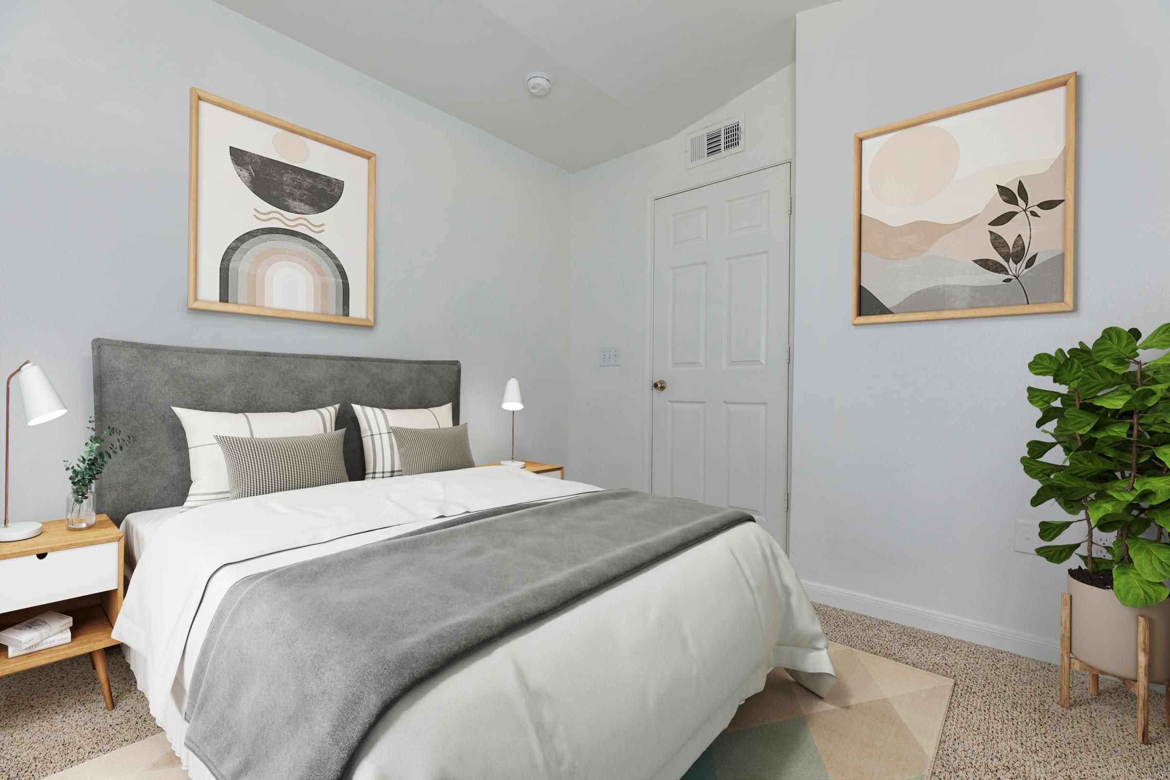 A cozy bedroom featuring a gray bedspread, two bedside lamps, and framed artwork on the walls. The room has light-colored walls, a door, and a potted plant in the corner, creating a calm and inviting atmosphere. The carpet is patterned with geometric shapes.