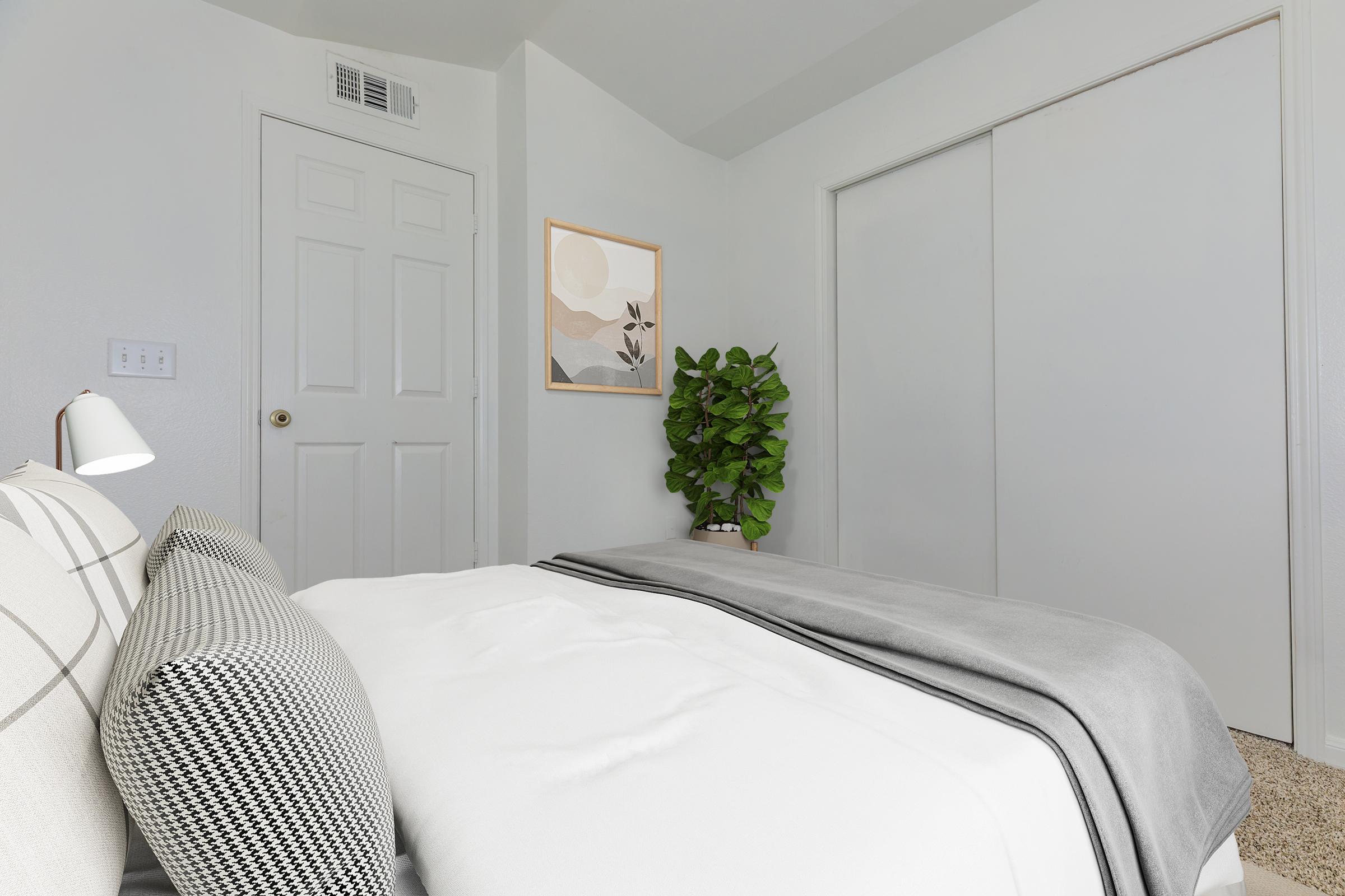 A cozy bedroom featuring a neatly made bed with a light blanket and decorative pillows. There's a small plant in the corner, a piece of wall art depicting abstract shapes, and a closet with sliding doors. The room has light-colored walls and a soft carpet, creating a serene atmosphere.