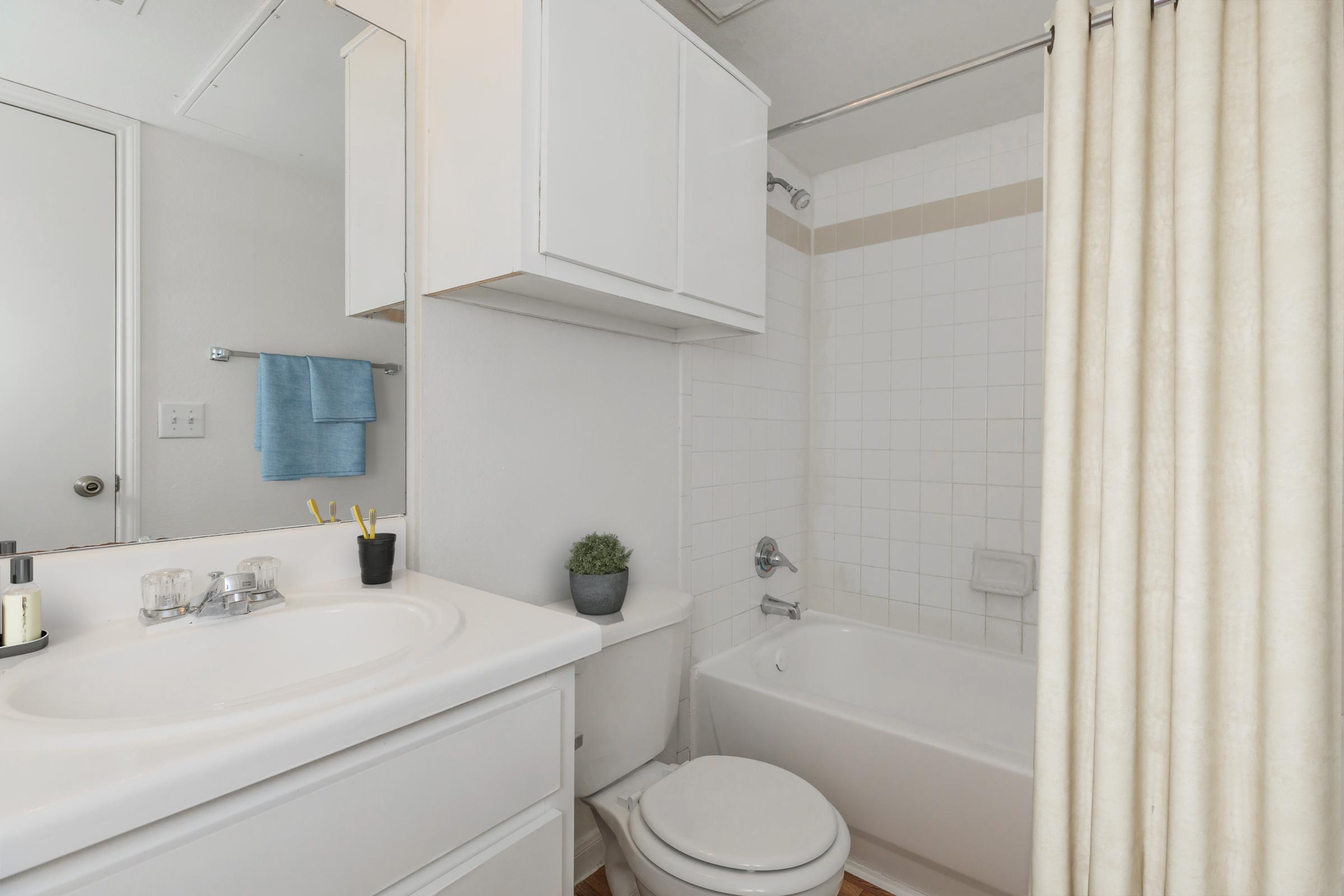 A clean, modern bathroom featuring a white sink with a mirror above, a bathtub with a shower curtain, and light-colored tiles. There is a small potted plant on the countertop and towels hanging nearby. The overall design is bright and functional, with a sleek look.