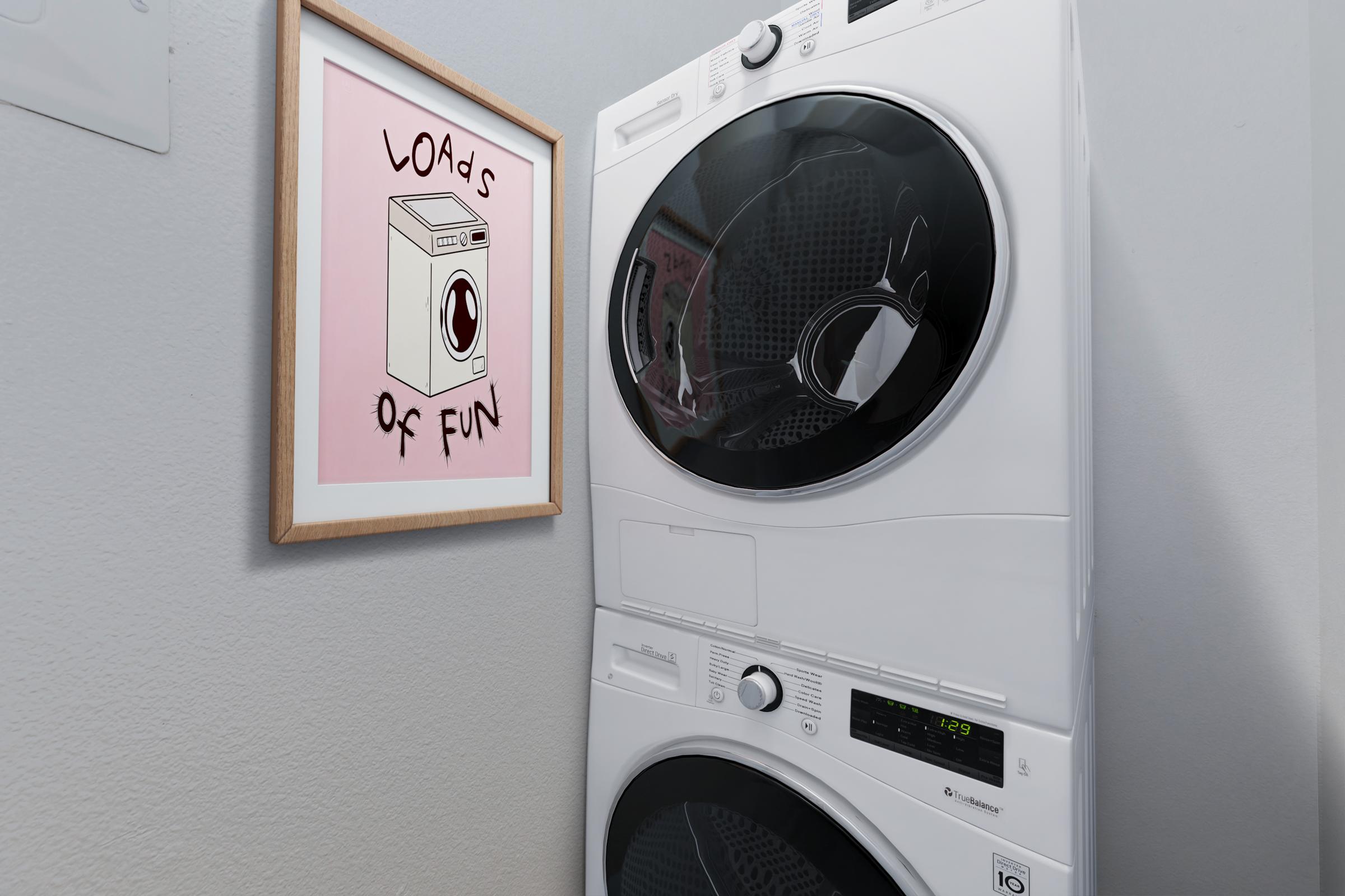 A stacked washer and dryer set in a laundry room, accompanied by a framed illustration on the wall that reads "Loads of Fun" with a whimsical depiction of a washing machine. The room has light gray walls and a clean, modern aesthetic.