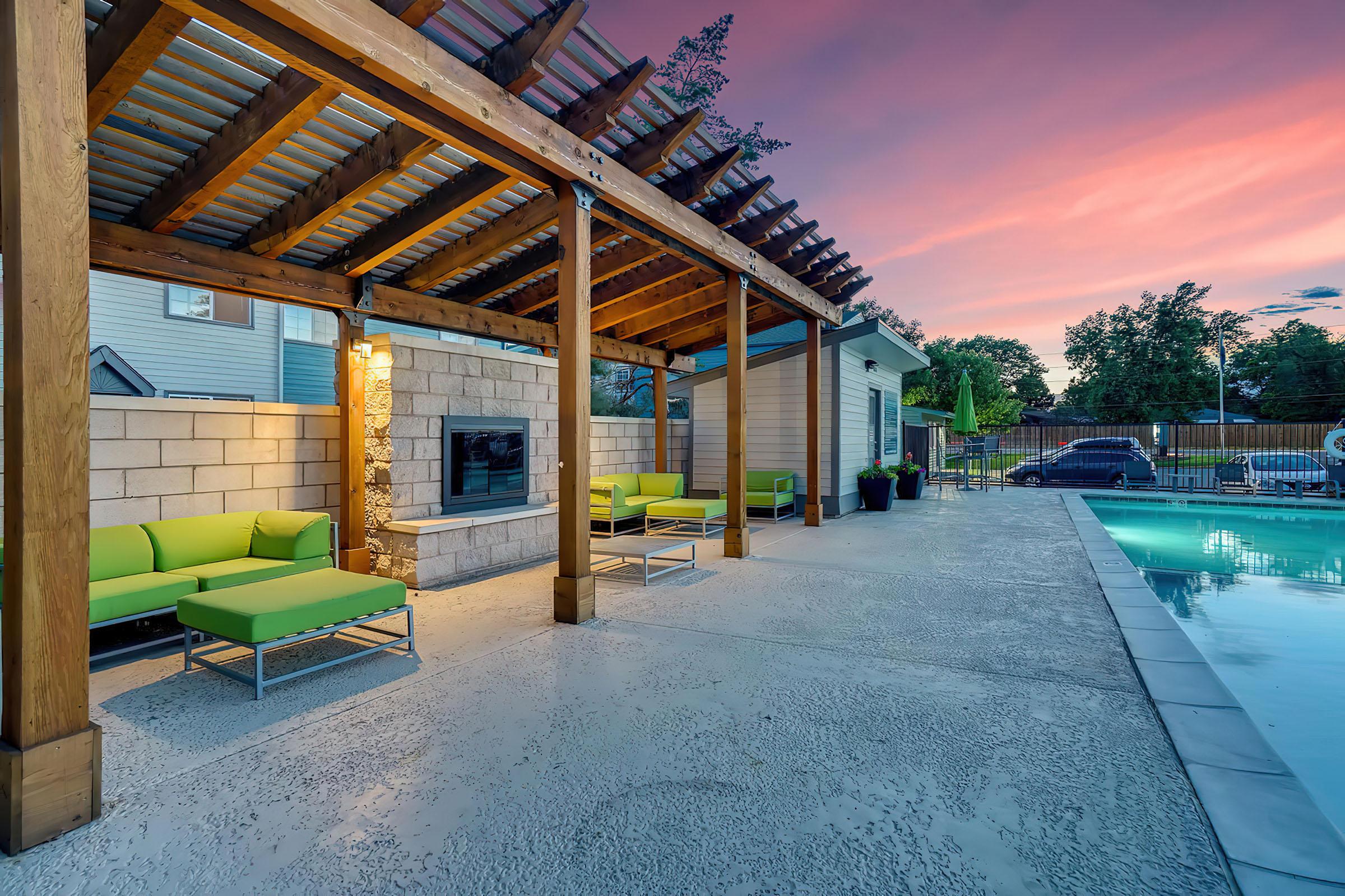 A modern outdoor space featuring green seating under a wooden pergola, with a wall-mounted fireplace. A swimming pool is visible nearby, surrounded by a paved area, and a colorful sunset sky adds ambiance to the scene.