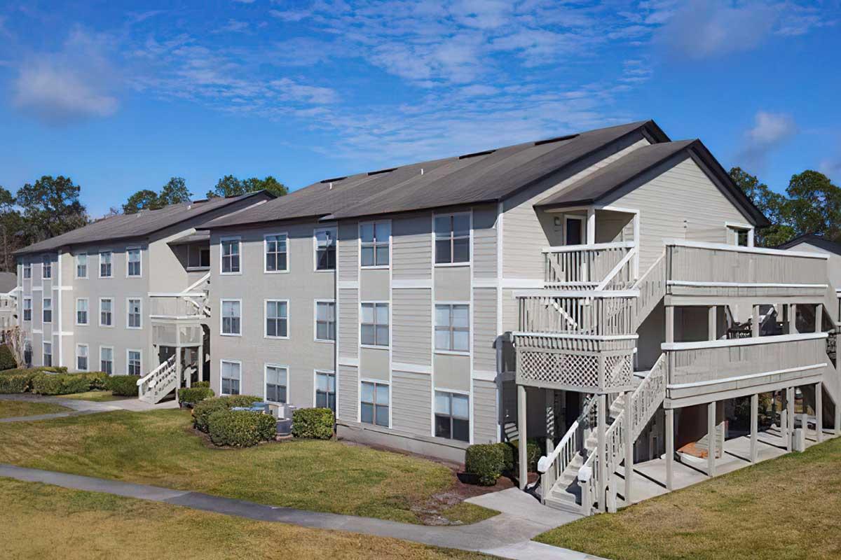 A multi-story apartment building with a light gray exterior, featuring balconies and a well-maintained lawn in the foreground. The sky is clear with a few clouds, creating a bright and inviting atmosphere.