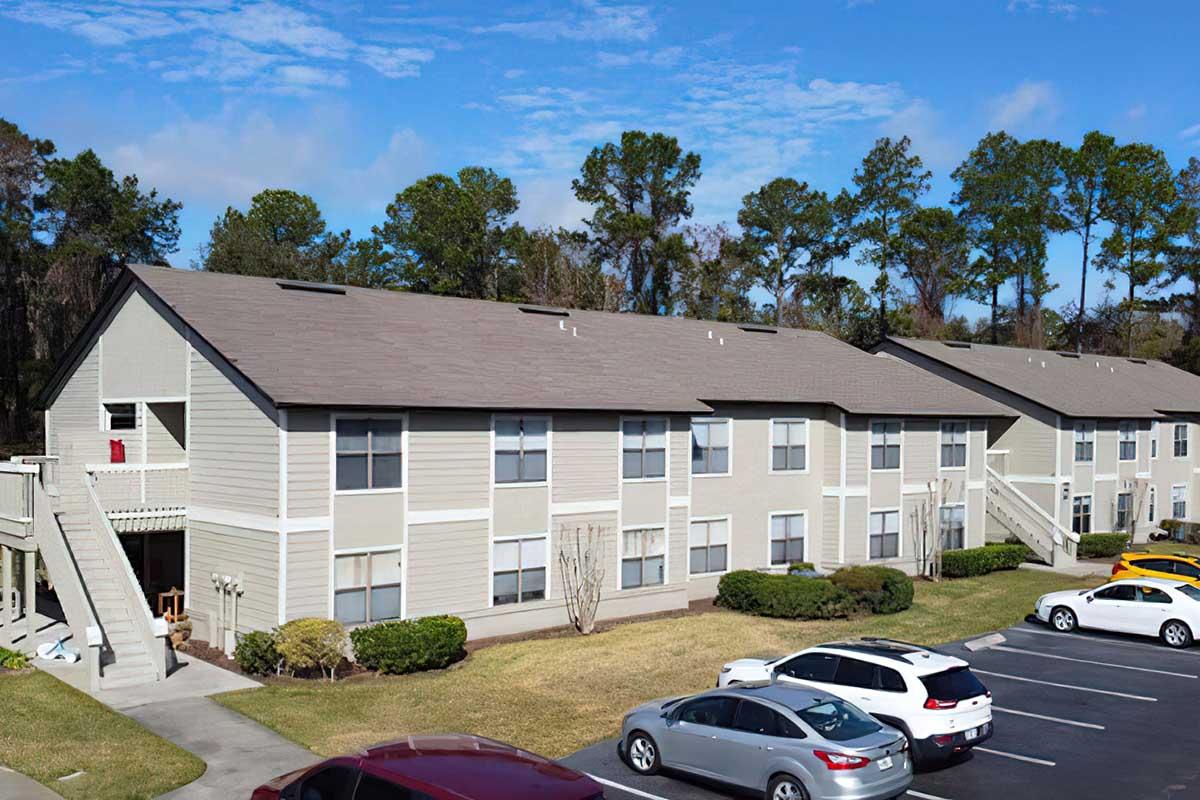 A row of two-story beige apartment buildings with balconies, surrounded by well-maintained grass and shrubs. Several parked cars are visible in the foreground, and tall trees are present in the background under a clear blue sky.