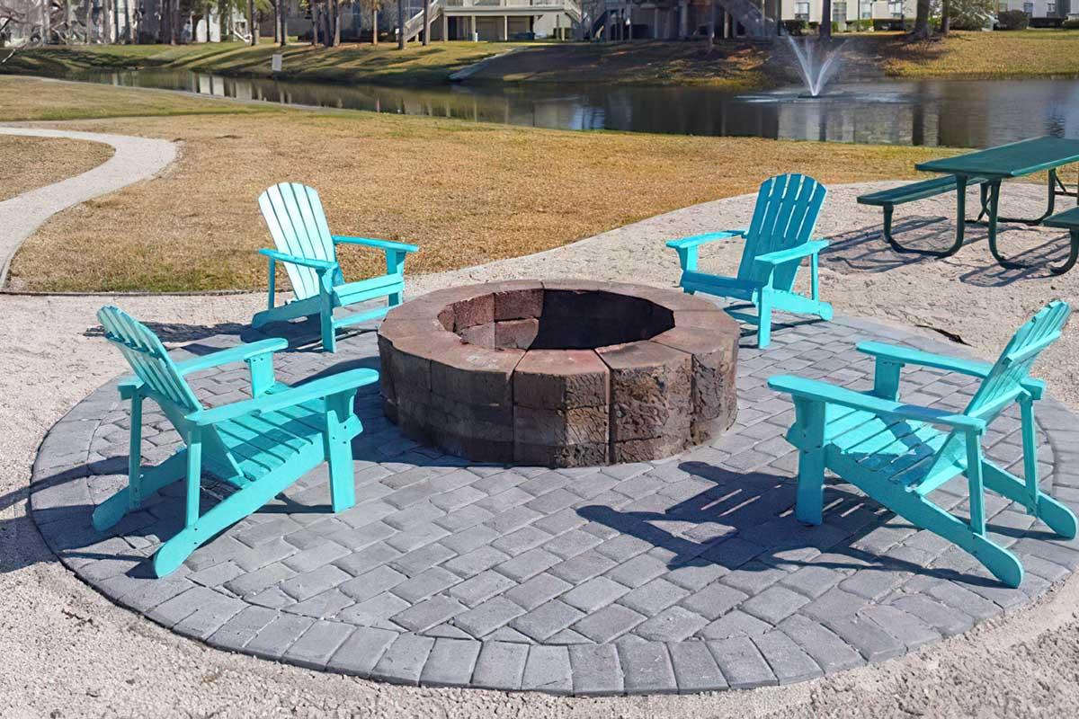A circular stone fire pit surrounded by four blue adirondack chairs, situated on a paved area near a grassy field and a pond. In the background, a picnic table can be seen, and a fountain is located in the pond. The scene is bright and inviting, perfect for gatherings.