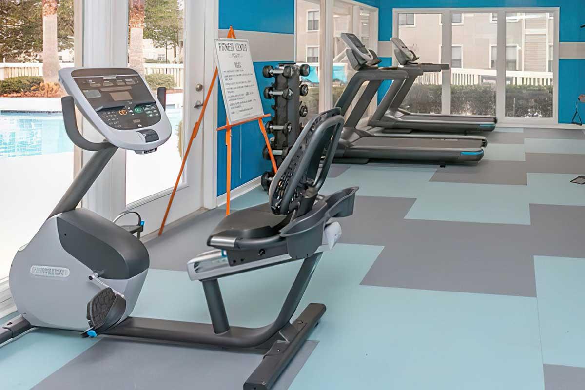 A fitness center featuring a stationary bike, an adjustable seated exercise bike, and several treadmills. The walls are painted in blue, and the flooring is a mix of light blue and gray tiles. A whiteboard with an easel is present, indicating it might provide additional information or instructions.