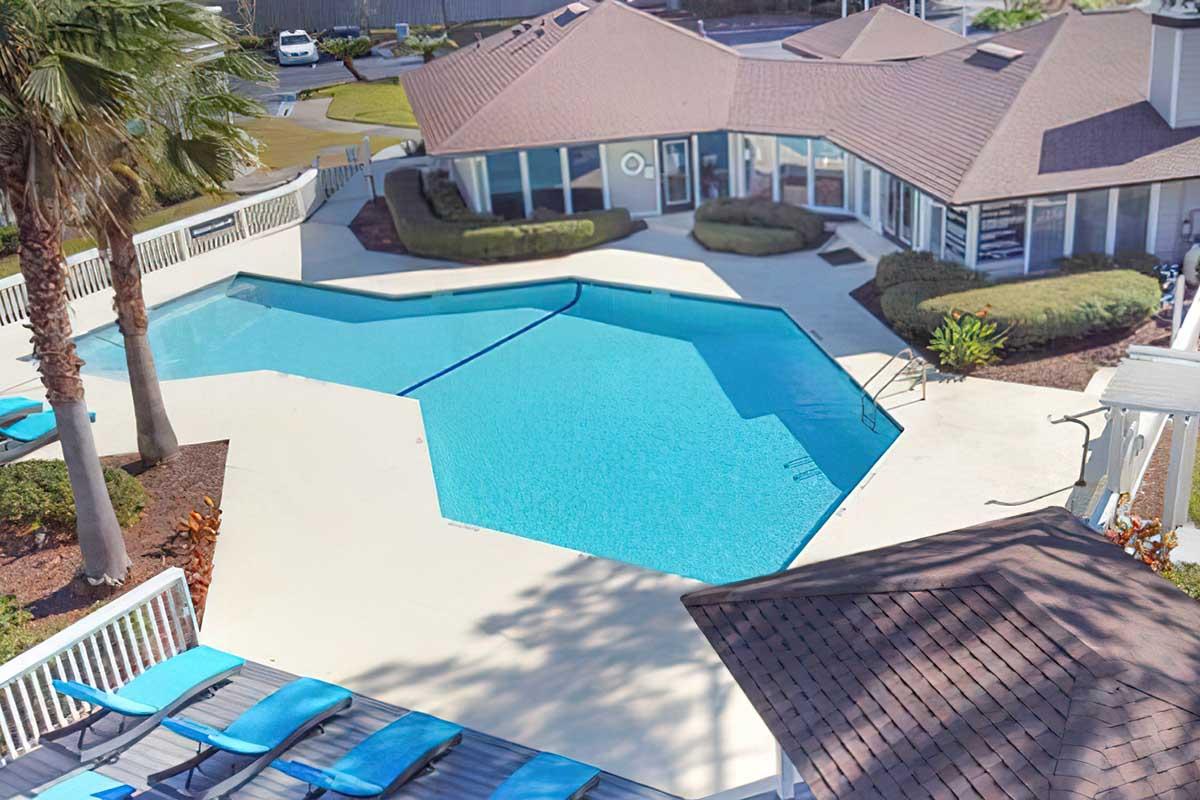 Aerial view of a geometric-shaped swimming pool surrounded by a deck with blue lounge chairs. The pool is clear and reflects sunlight, with landscaped areas featuring palm trees and shrubs. The nearby building has large windows and a sloped roof, indicating a relaxing outdoor space.