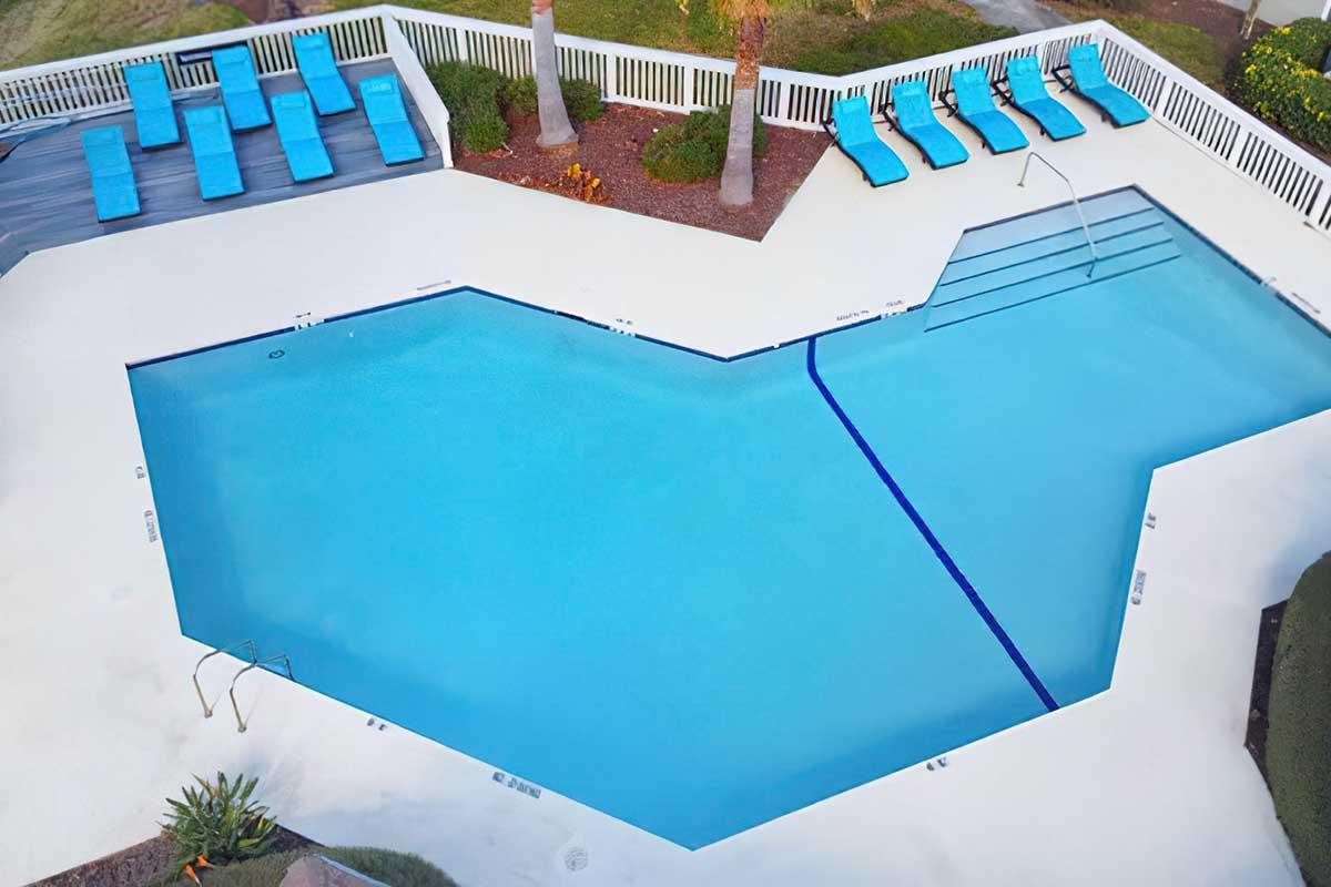 A heart-shaped swimming pool surrounded by lounge chairs and palm trees, featuring a white deck and landscaped greenery. The pool is clear blue, with steps leading in, and the area is well-maintained and inviting.